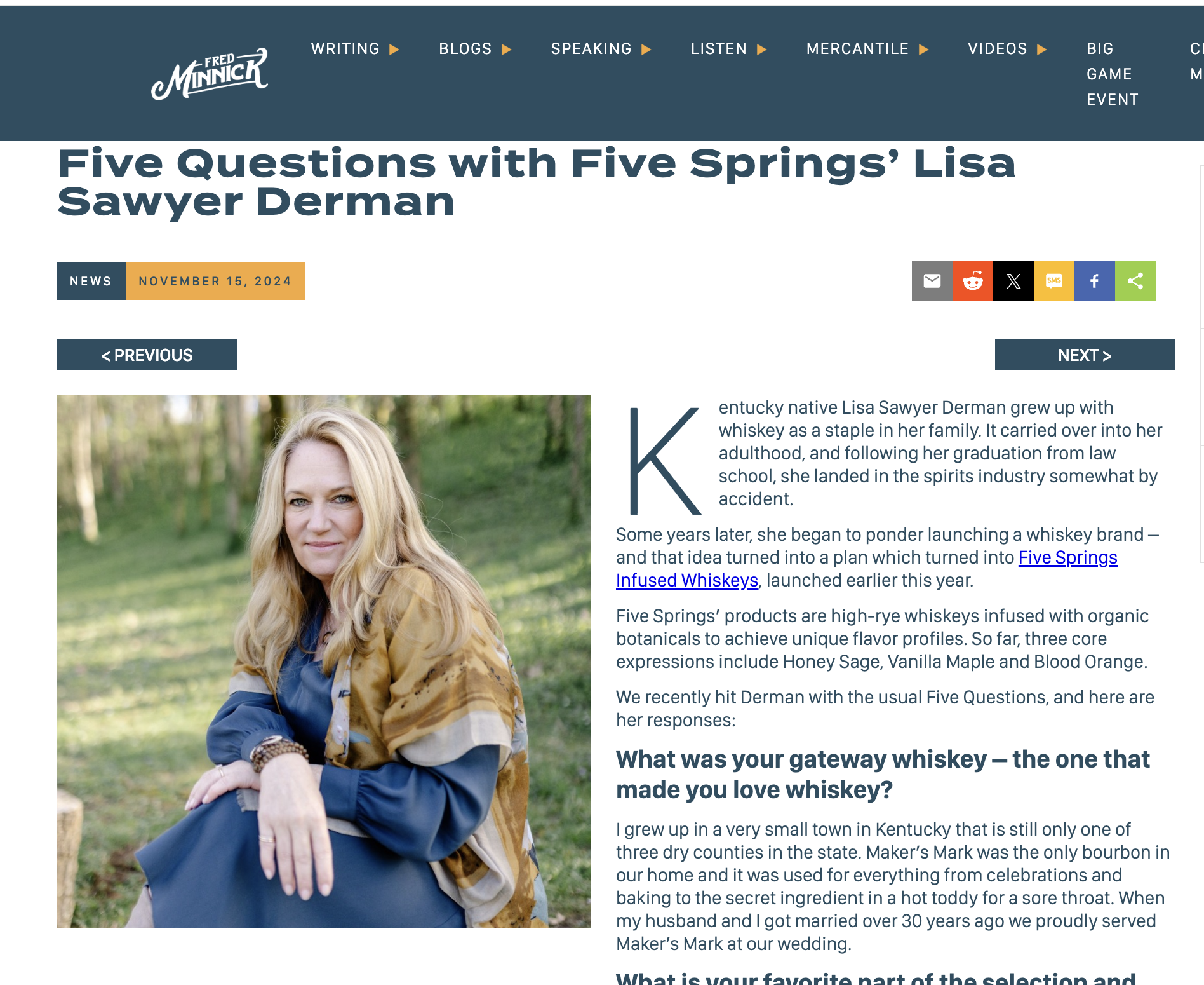 A woman with long blonde hair sitting outdoors on a grassy area with trees in the background, wearing a blue dress and a patterned shawl.