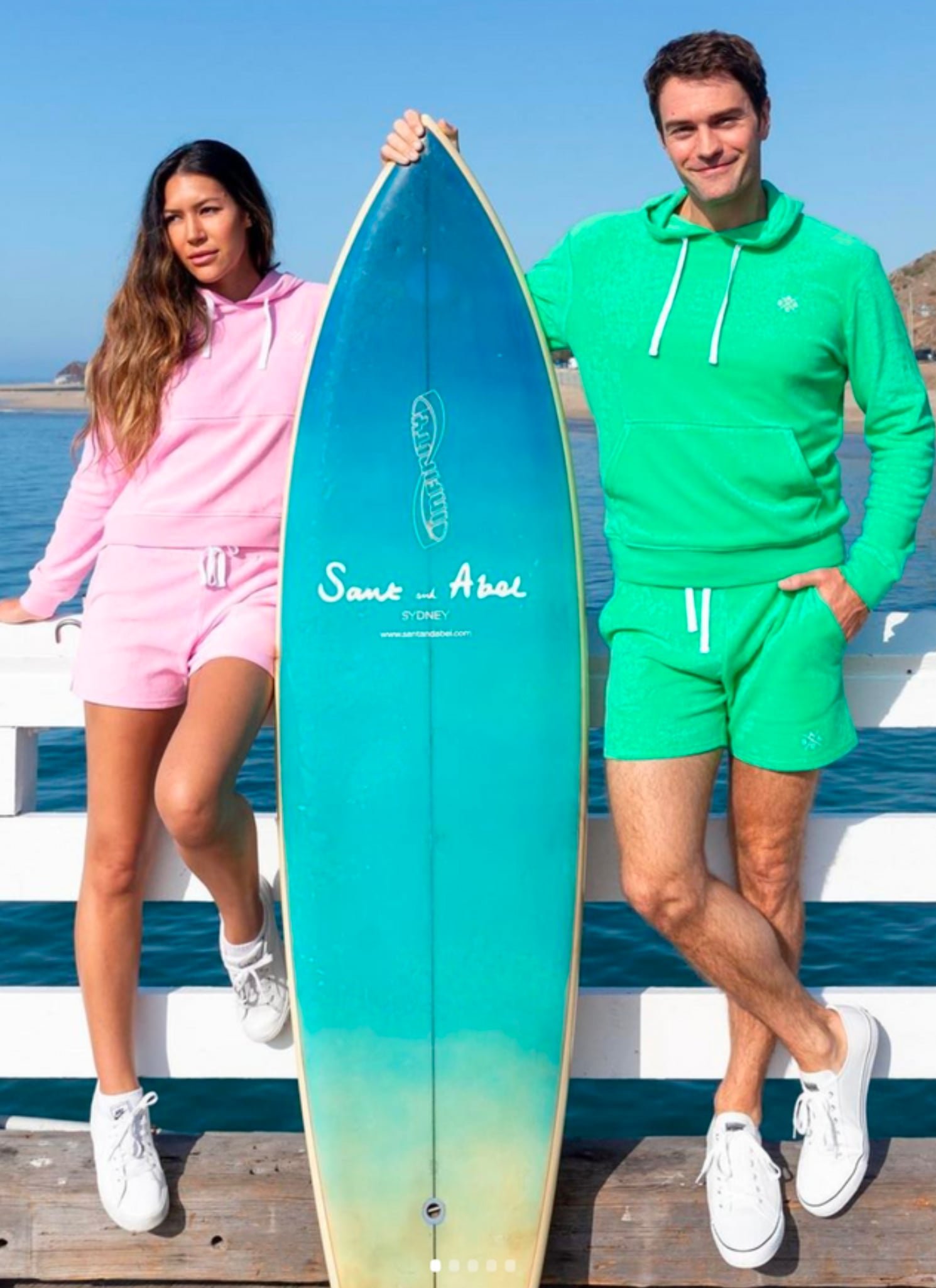 man and woman in bright colored clothing by the beach with a surf board