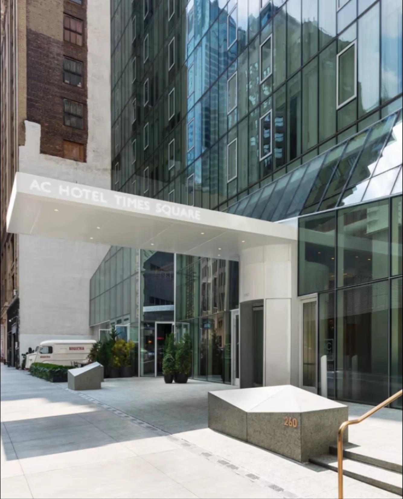Modern glass entrance of AC Hotel Times Square with street view, potted plants, and reflections of buildings on the windows.