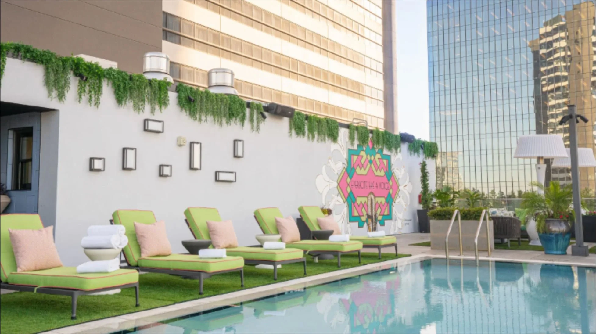 Rooftop pool with green lounge chairs, rolled towels, and city buildings in the background on a sunny day.