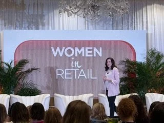 Woman speaking at a conference on women in retail, with a stage decorated with plants and a large screen background.