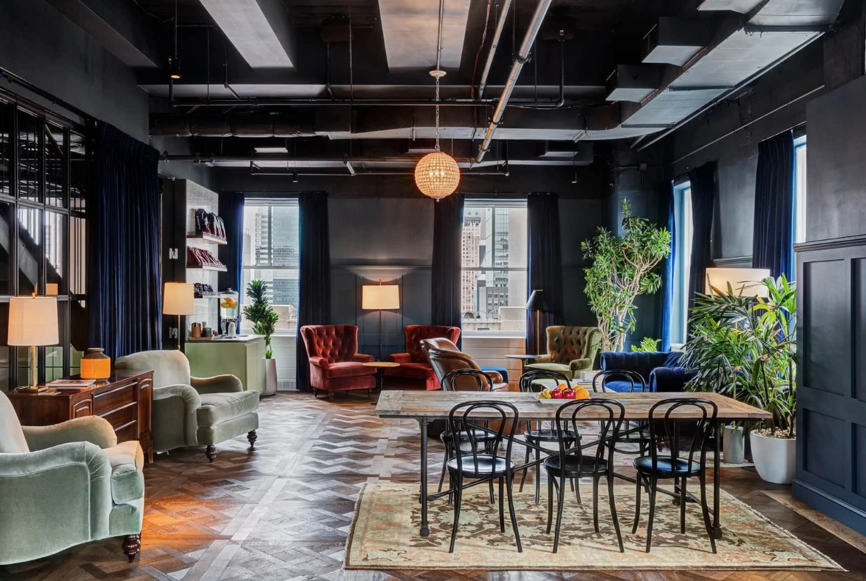 Chic lounge with velvet armchairs, dining table, green plants, large windows, and modern lighting in a stylish, dark-toned interior.
