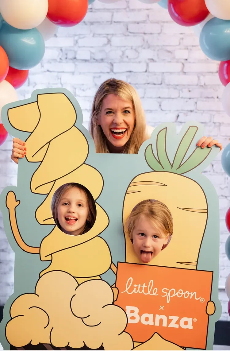 A woman with two children behind a cartoon-style cutout of a large carrot and a bunch of pasta, with balloons in the background.