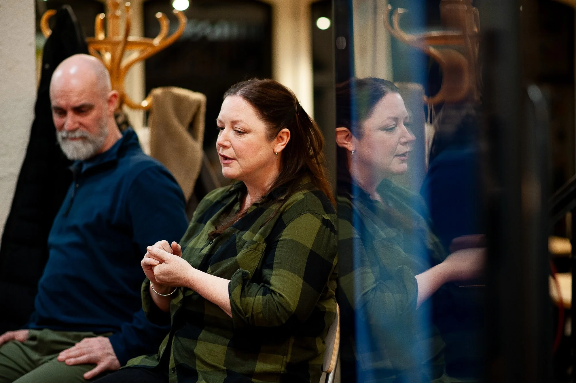 A woman with brown hair praying with her hands closed in front of her eyes, sitting next to a man with a bald head with a gray beard, both seated in a room with a coat rack and a mirror or glass panel reflecting her image.