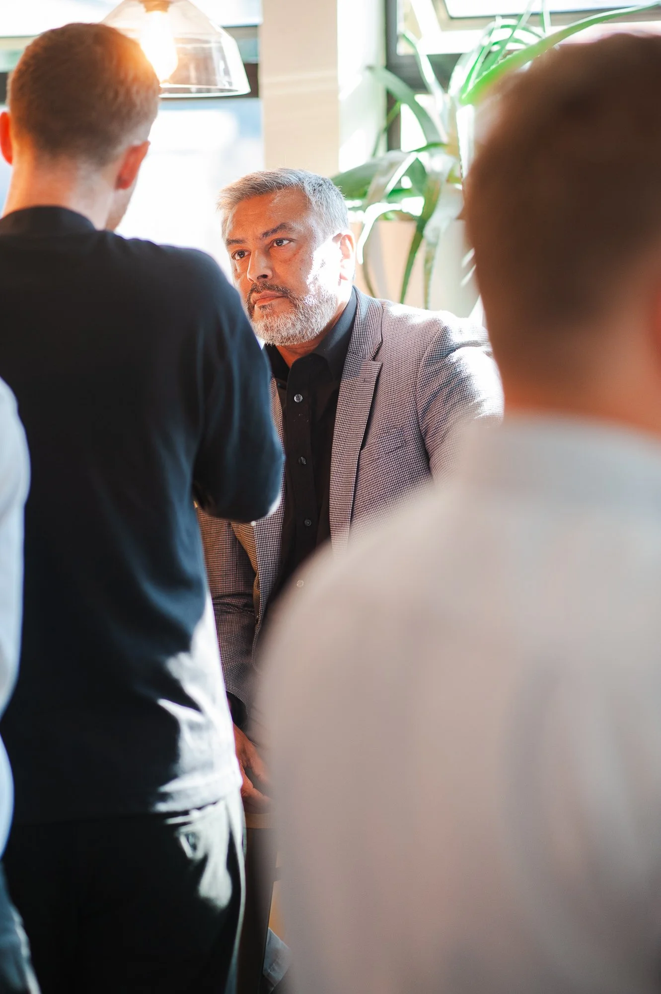 A group of four men having a discussion indoors. The man in the center has gray hair and a beard, wearing a gray blazer and black shirt, sitting at a table near a window with sunlight and greenery nearby.