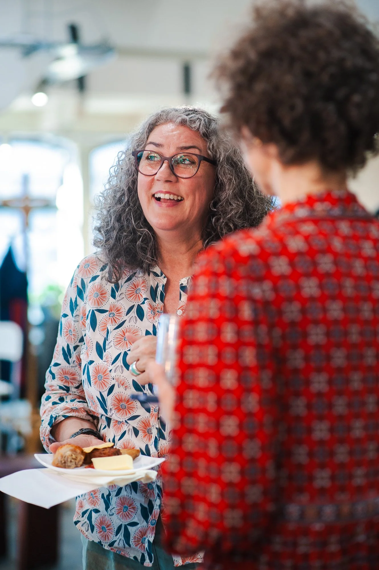 A woman with gray curly hair and glasses talking to a person in a red patterned shirt, holding a plate of food in her hand.