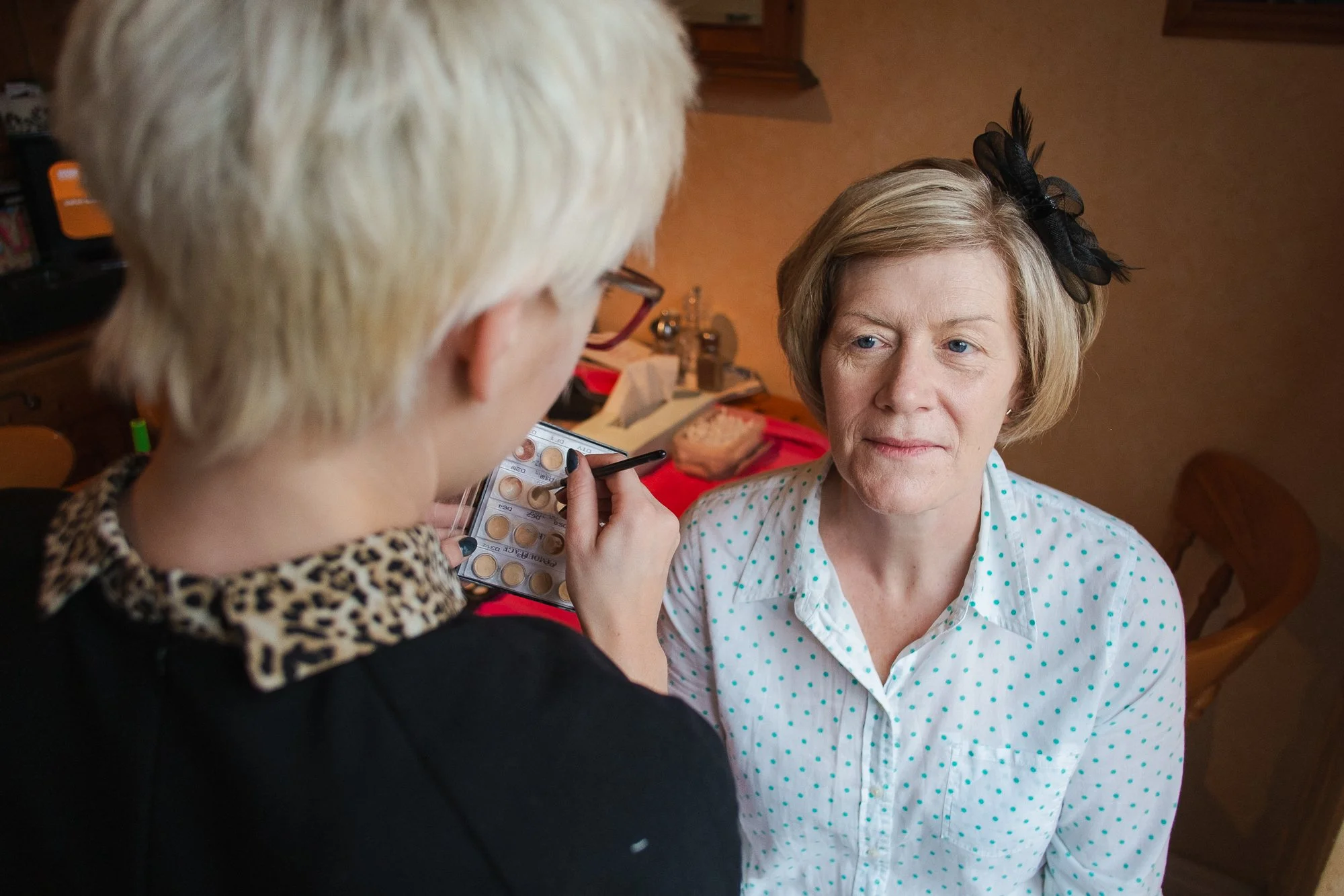 A woman having makeup applied by a person with short blond hair, indoors with warm lighting. The woman has short blonde hair with a black bow and wears a white shirt with blue polka dots.