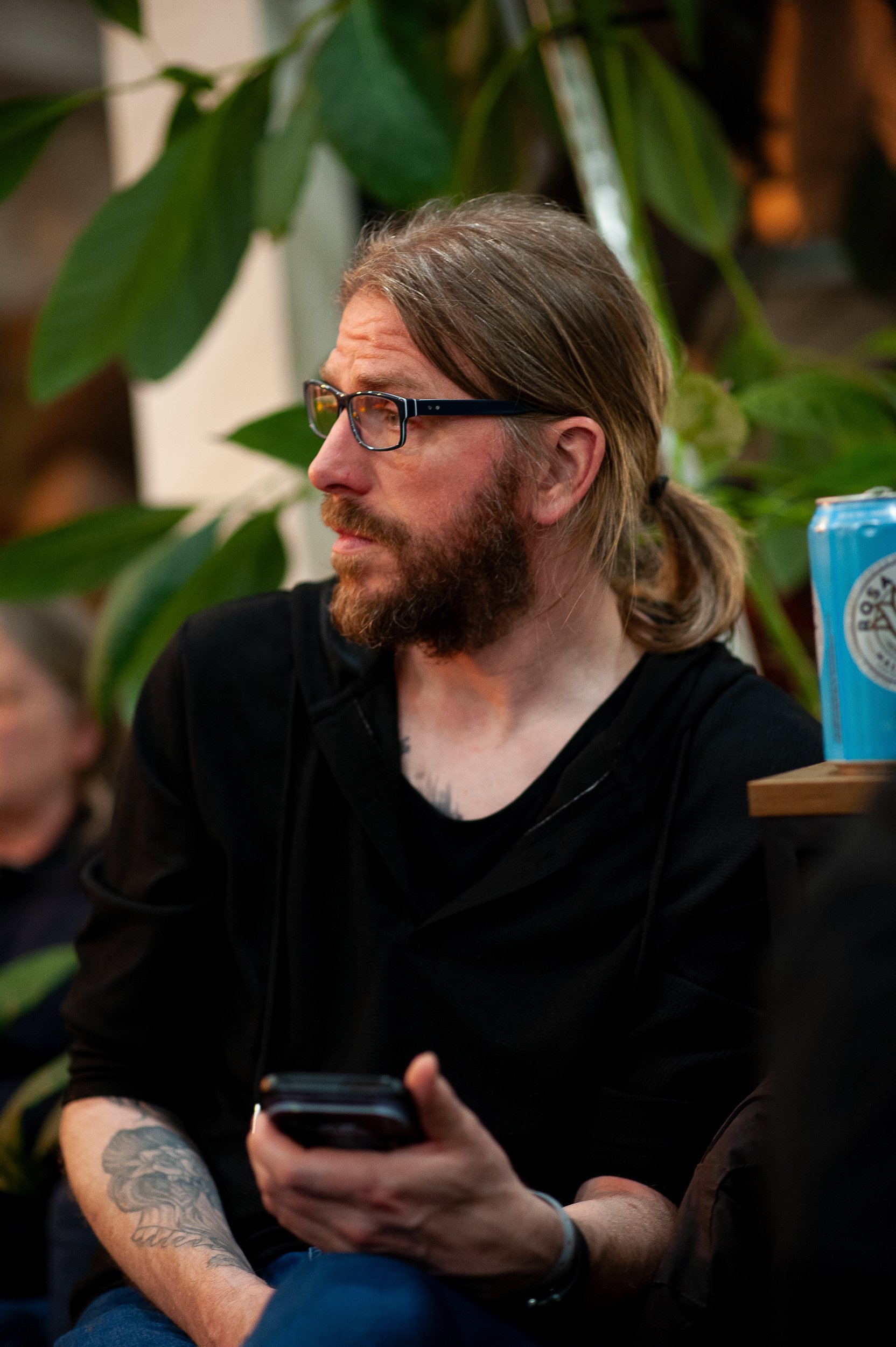 Man with glasses and ponytail holding a smartphone, surrounded by green plants, with a blue can and tattoo on his right arm.
