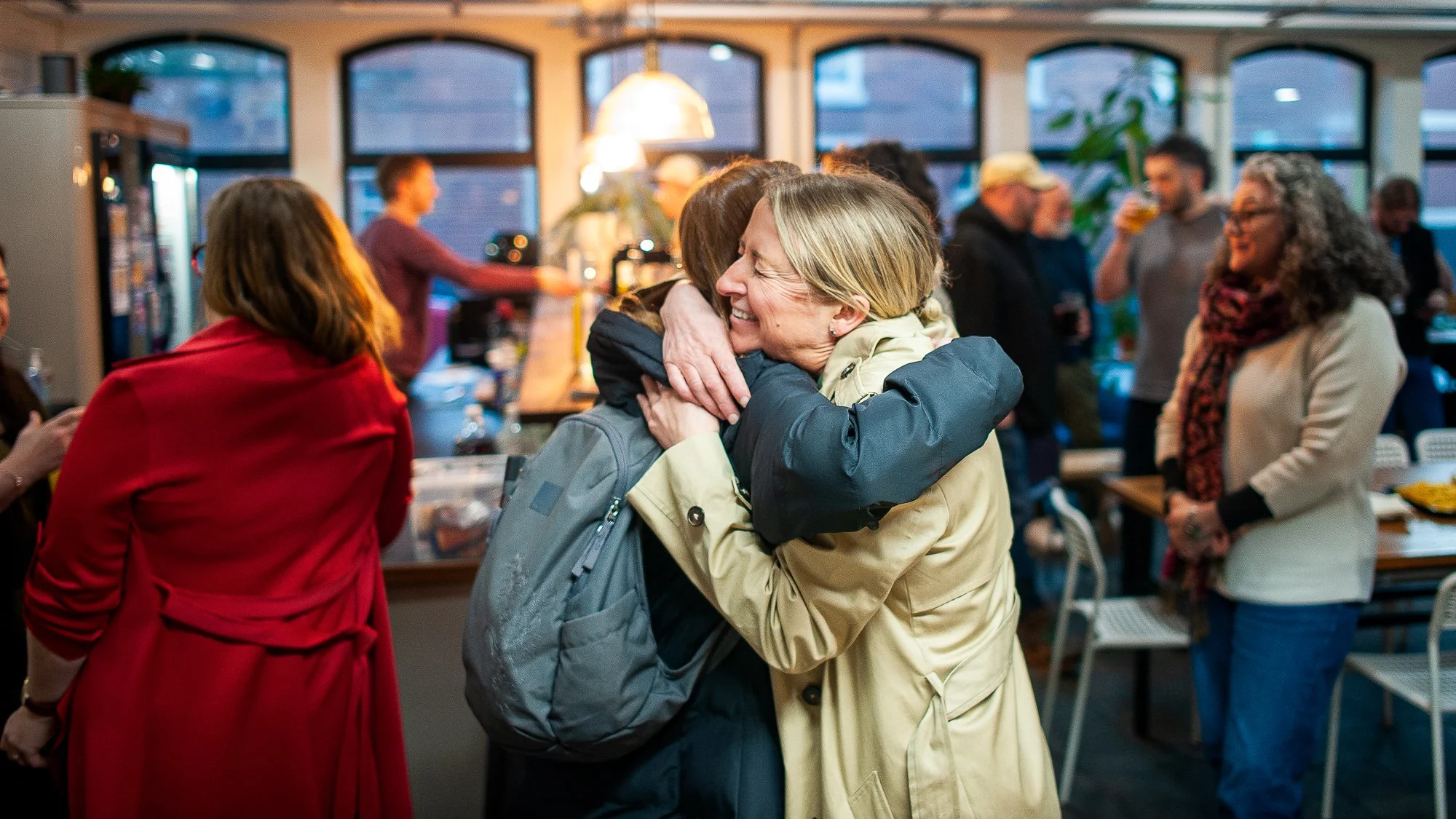 Two women hugging and smiling at a social gathering in a brightly lit room with large windows and several other people in the background.