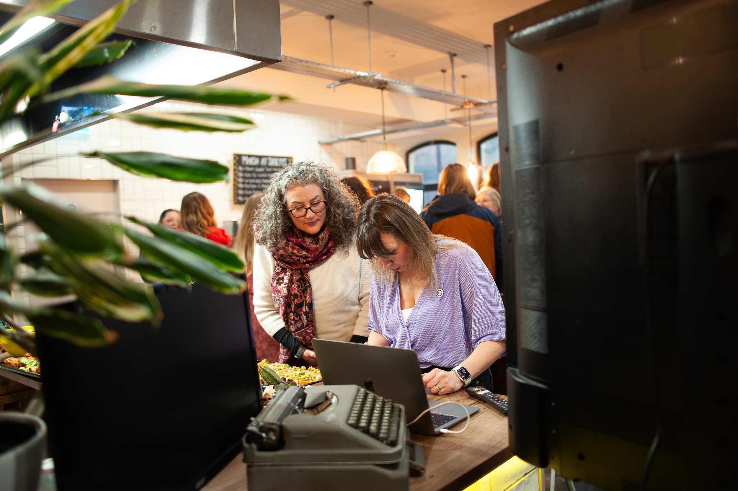 Two women in a kitchen, one using a laptop, with a typewriter and potted plant nearby. Several people are in the background.
