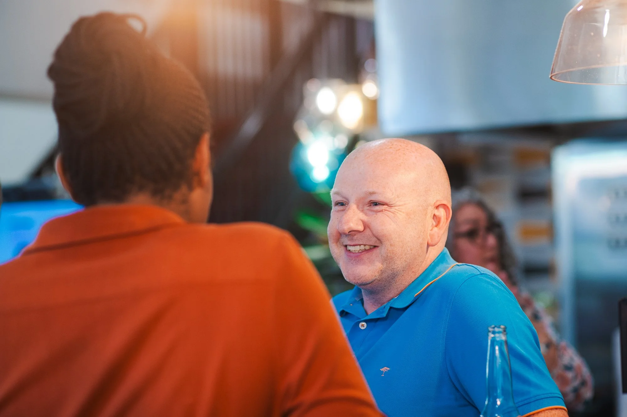 Smiling man in a blue polo shirt talking to another person, with a woman in glasses in the background, in a lively indoor setting with warm lighting.