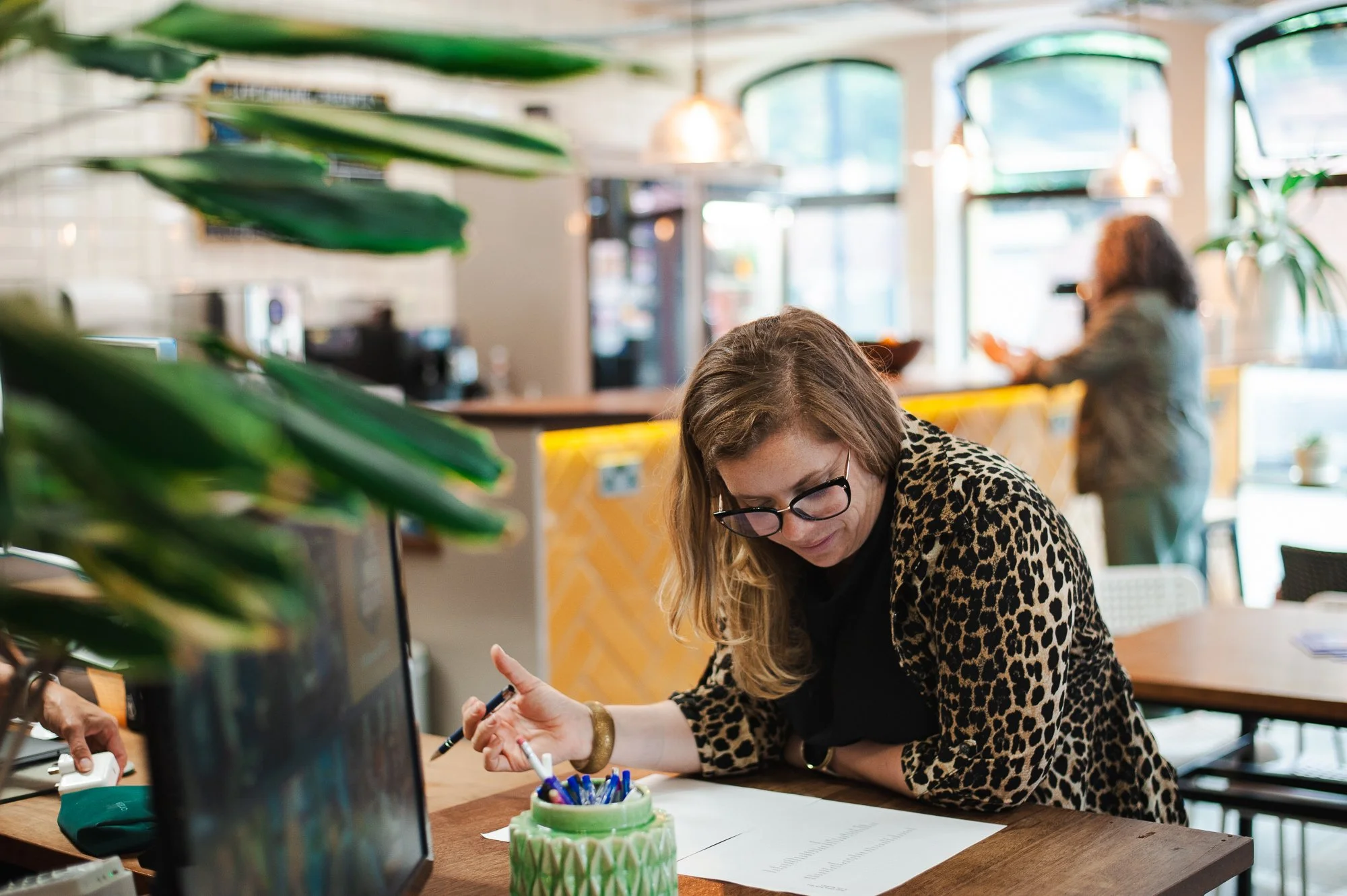 A woman with glasses and a leopard print jacket is at a cafe or restaurant, leaning on a wooden table with papers and a pen, while a person in the background is at the counter. The setting is well-lit with natural light and decorated with plants.