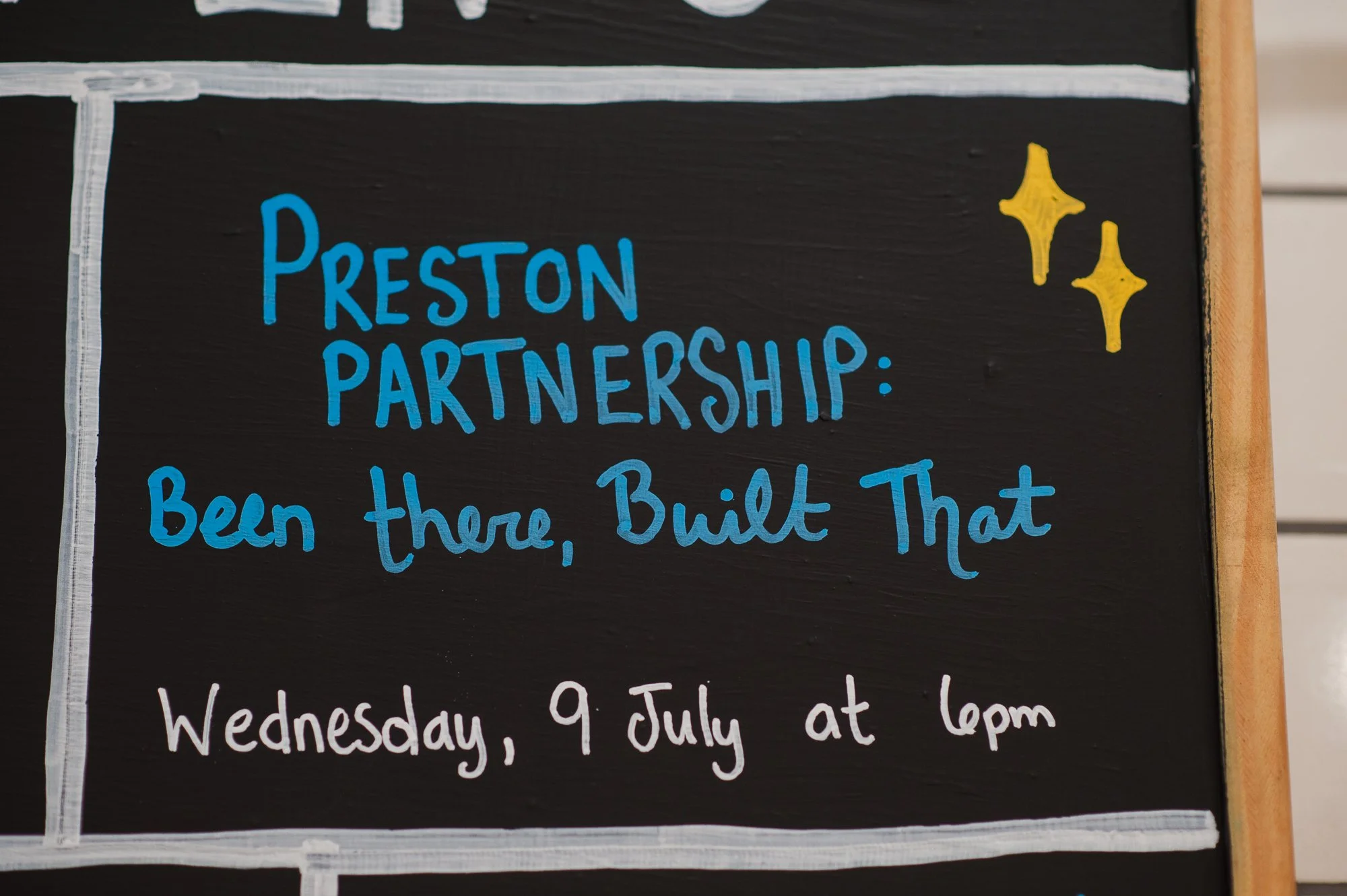 A blackboard sign with colorful handwritten text promoting a Preston Partnership event on July 9th, with decorative yellow stars.