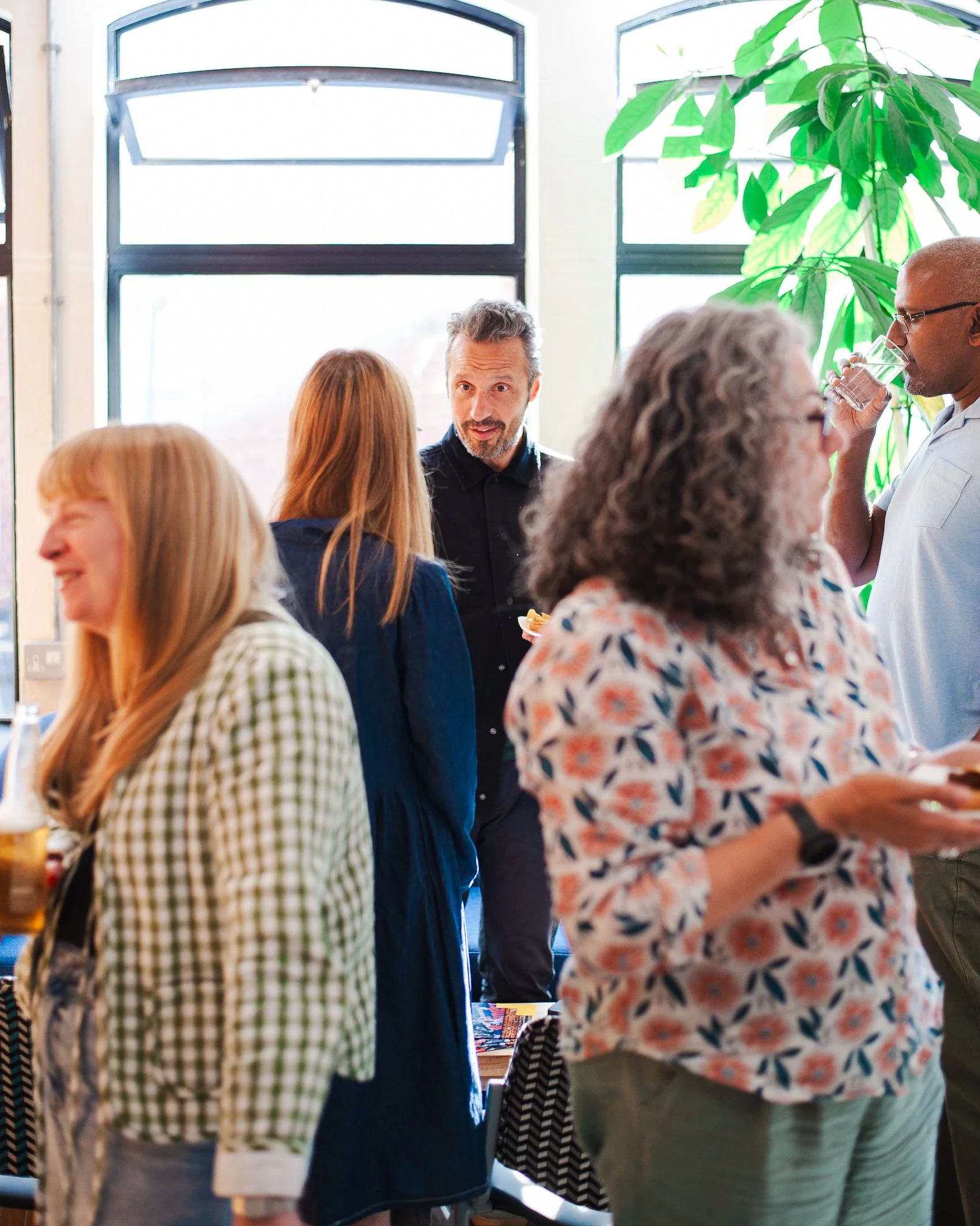 A group of people socializing at a gathering in a bright indoor space with large windows and a green plant.
