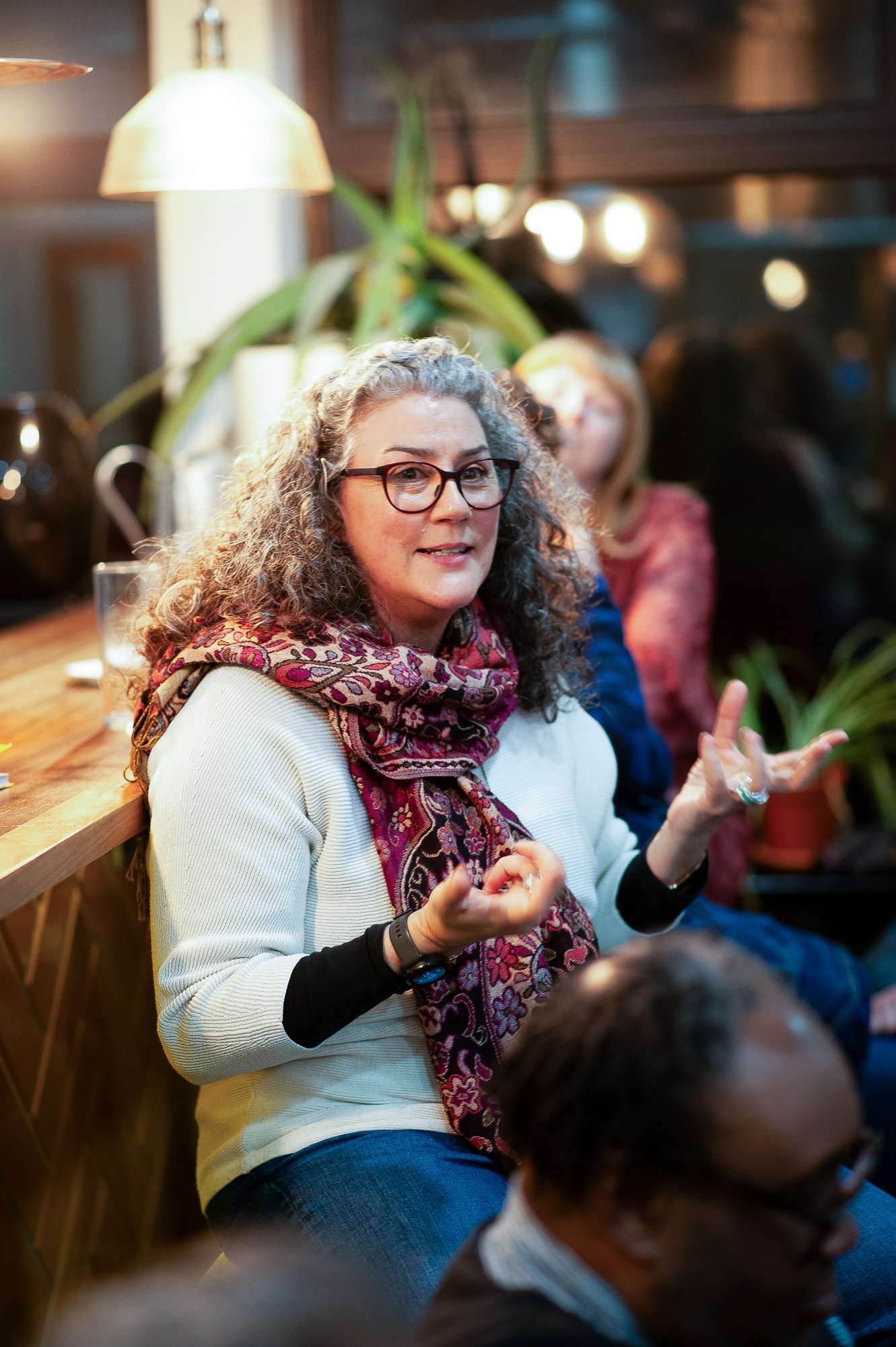A woman with curly gray hair, glasses, and a colorful scarf, speaking and making hand gestures during a discussion at an indoor gathering or event.