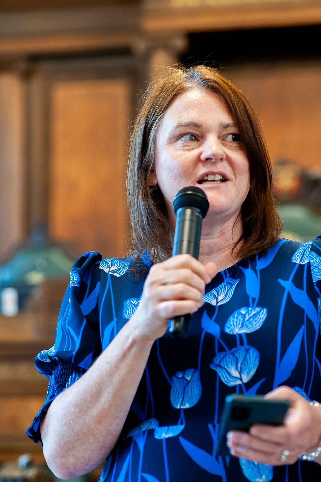A woman with brown hair wearing a blue floral dress speaking into a microphone and holding a phone in her other hand during a presentation or event.
