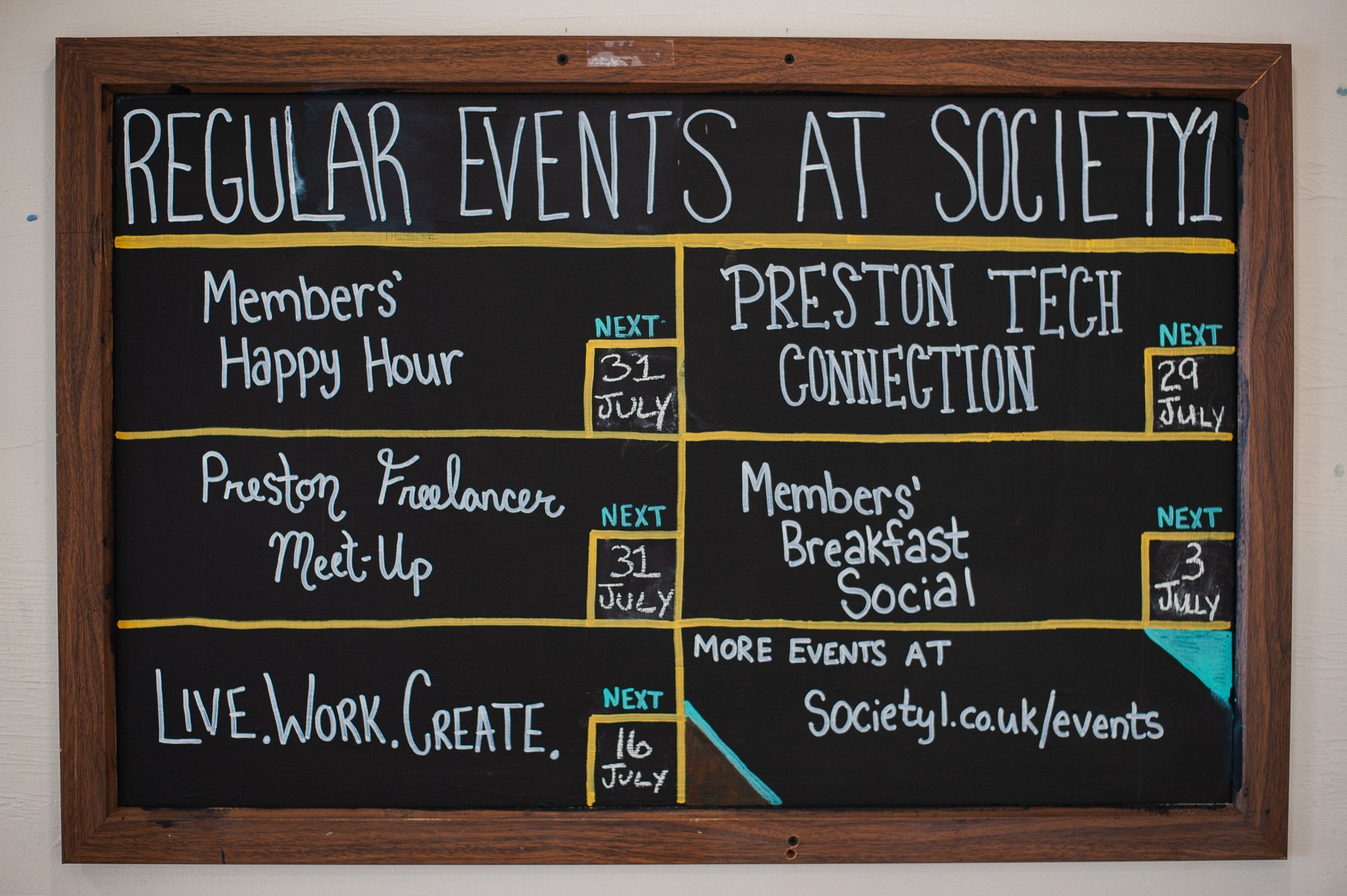 A chalkboard sign listing upcoming regular events at Society1, including Members' Happy Hour on July 31, Preston Freelancer Meet-Up on July 31, Members' Breakfast Social on July 3, and LIVE.WORK.CREATE on July 16, with additional events at society1.c