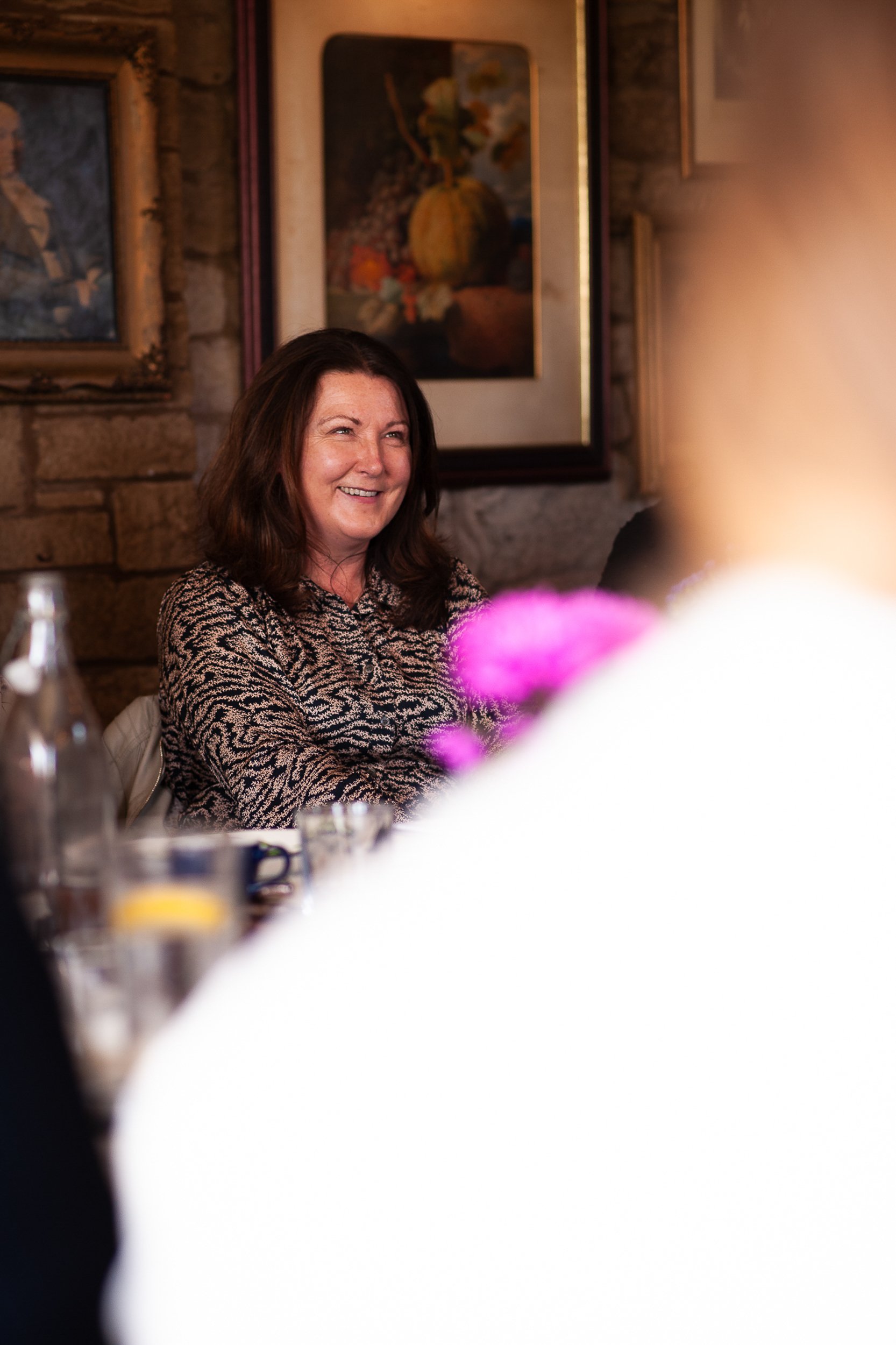Woman smiling while seated at a table in a cozy room with paintings on the wall.