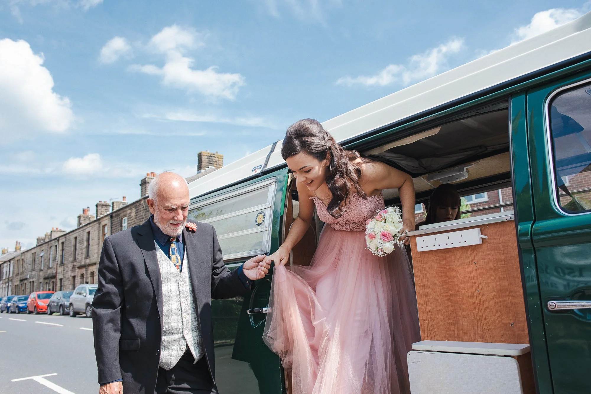 A bridesmaid in a pink dress stepping out of a green vintage van and being helped by an older man in a suit.