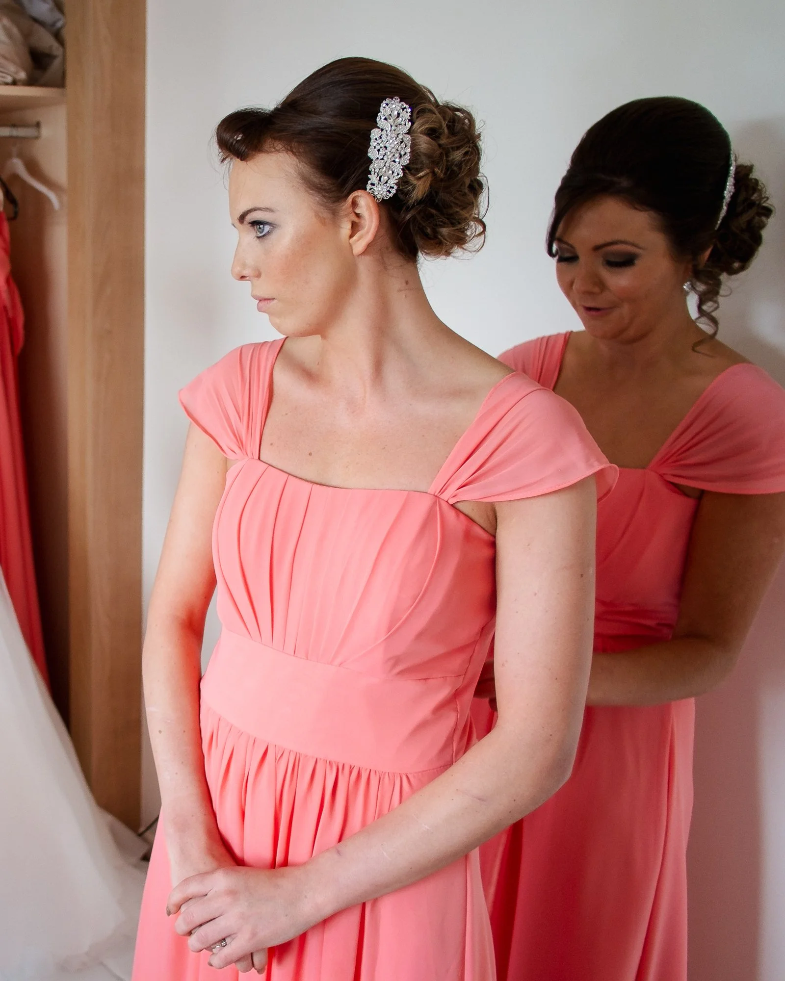 Brunette woman in coral dress with hair adorned with sparkly clip, standing beside another woman in similar dress
