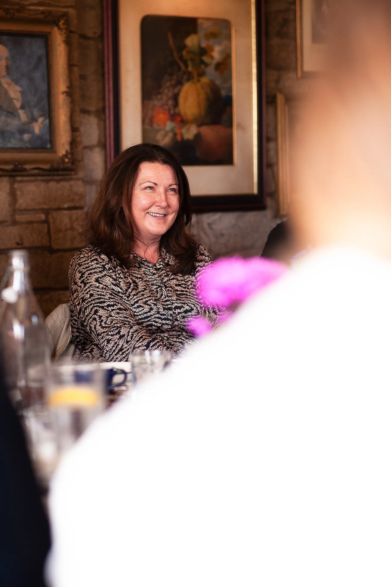 A woman with brown hair smiling and sitting at a table, with several paintings on a stone wall behind her. Blurred objects and flowers partially obscure the foreground.
