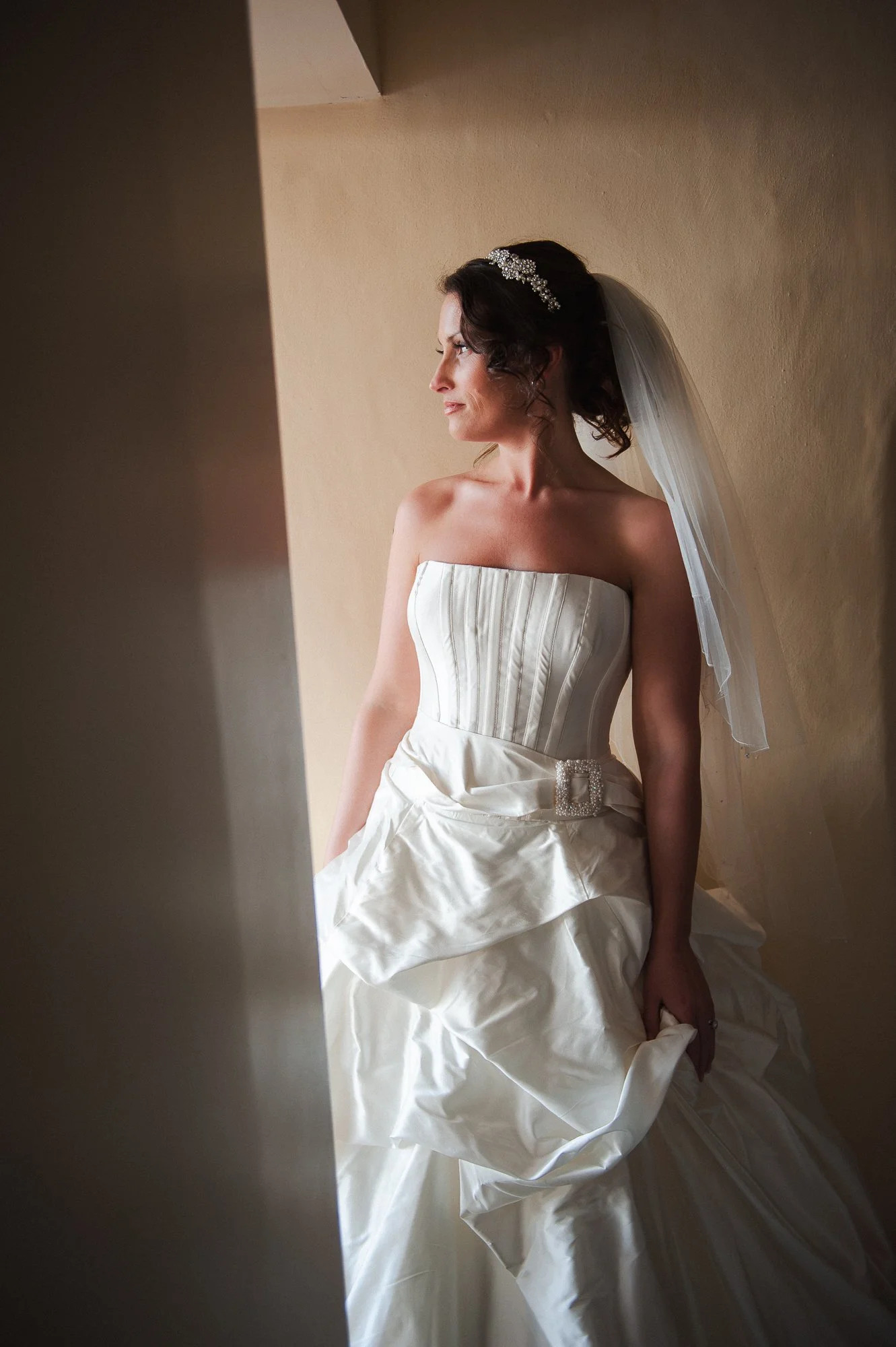 Bride in a strapless white wedding gown with a beaded belt, standing by a wall, looking away, wearing a veil and a hair accessory.