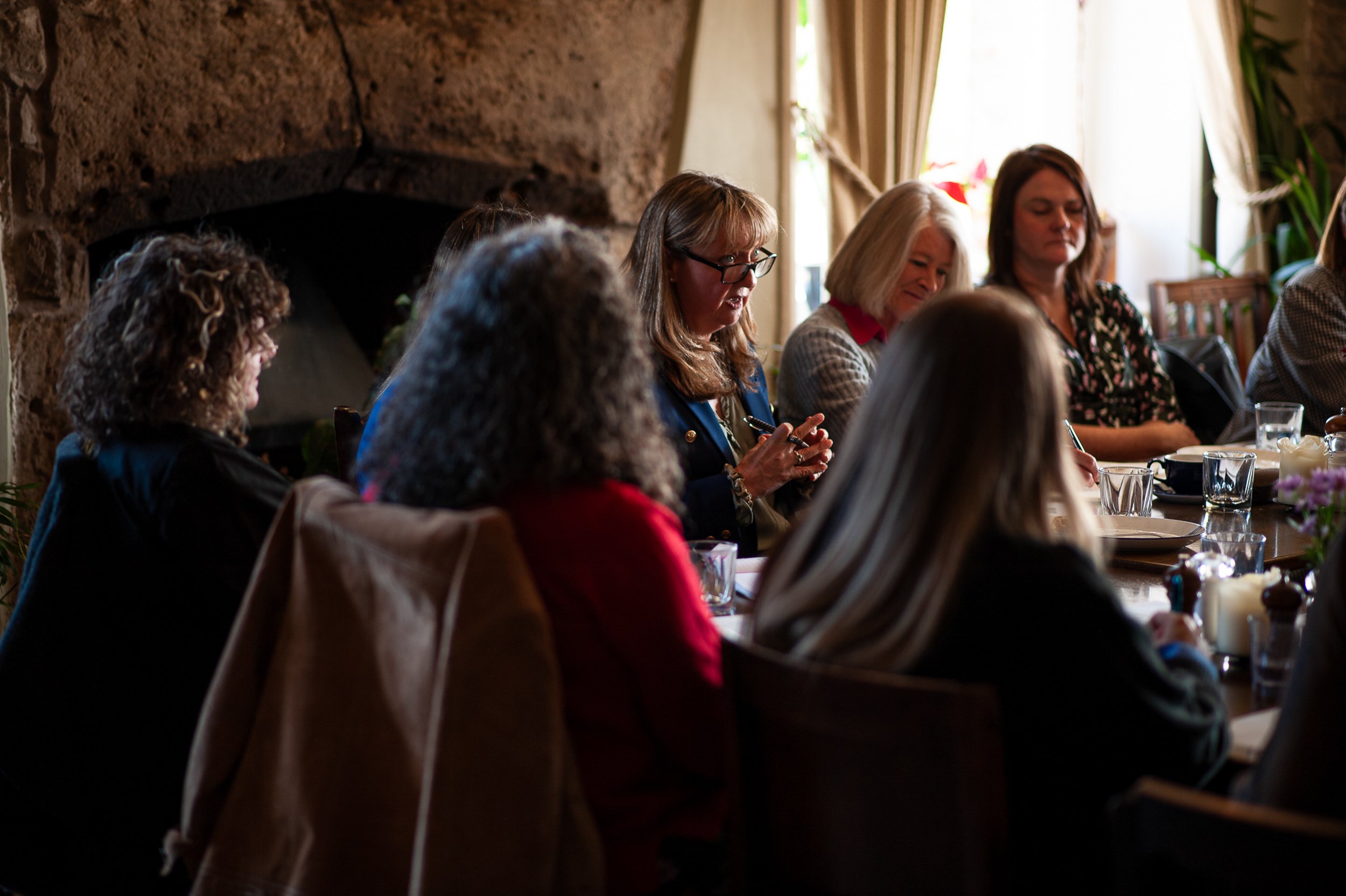 Group of women seated around a table, engaging in discussion, with one woman speaking. The setting is warmly lit, with a stone fireplace in the background and curtains framing a window.