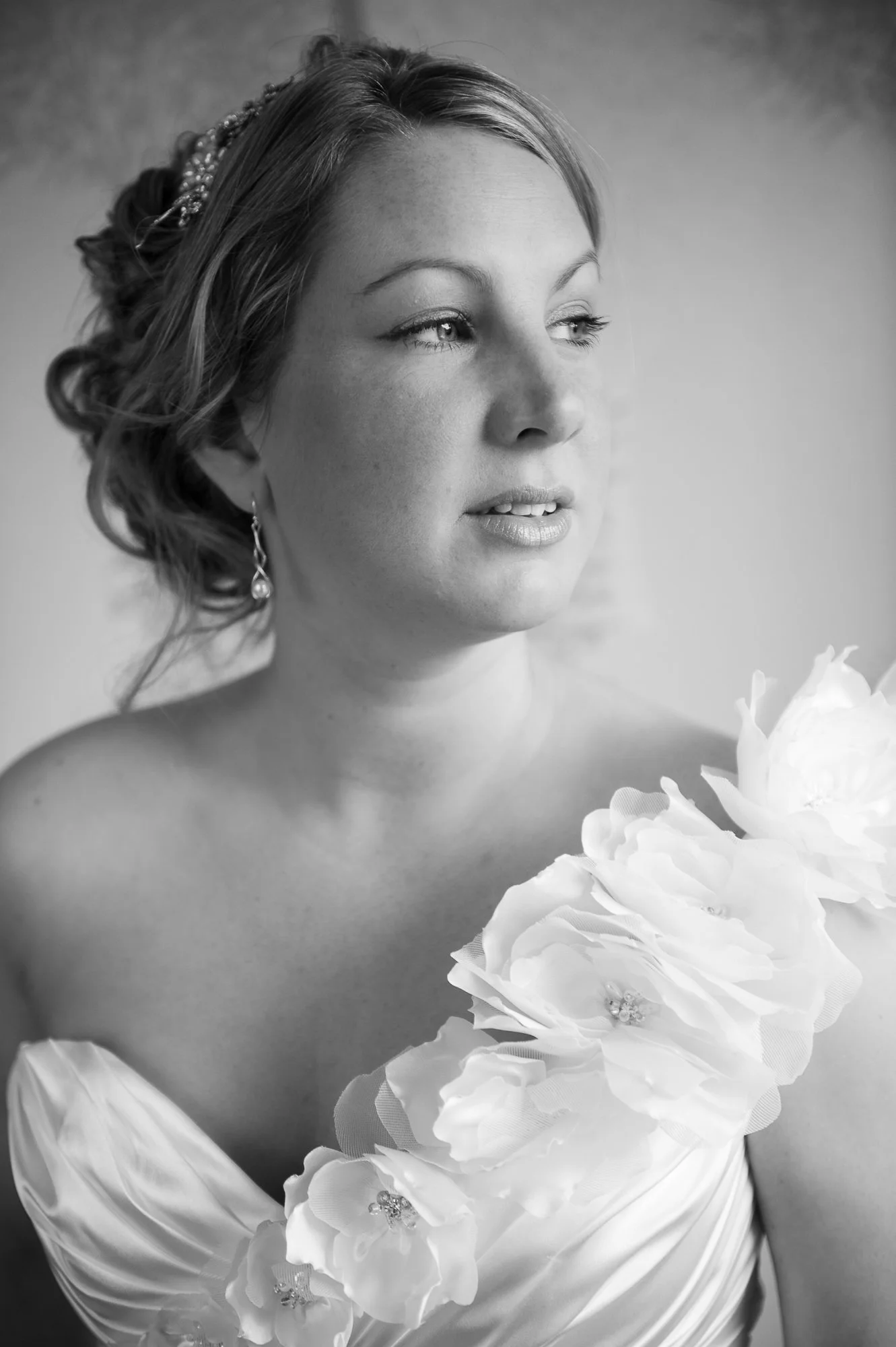 A black-and-white portrait of a bride with styled hair, wearing earrings and a dress with large floral embellishments on the shoulder, looking to the side.