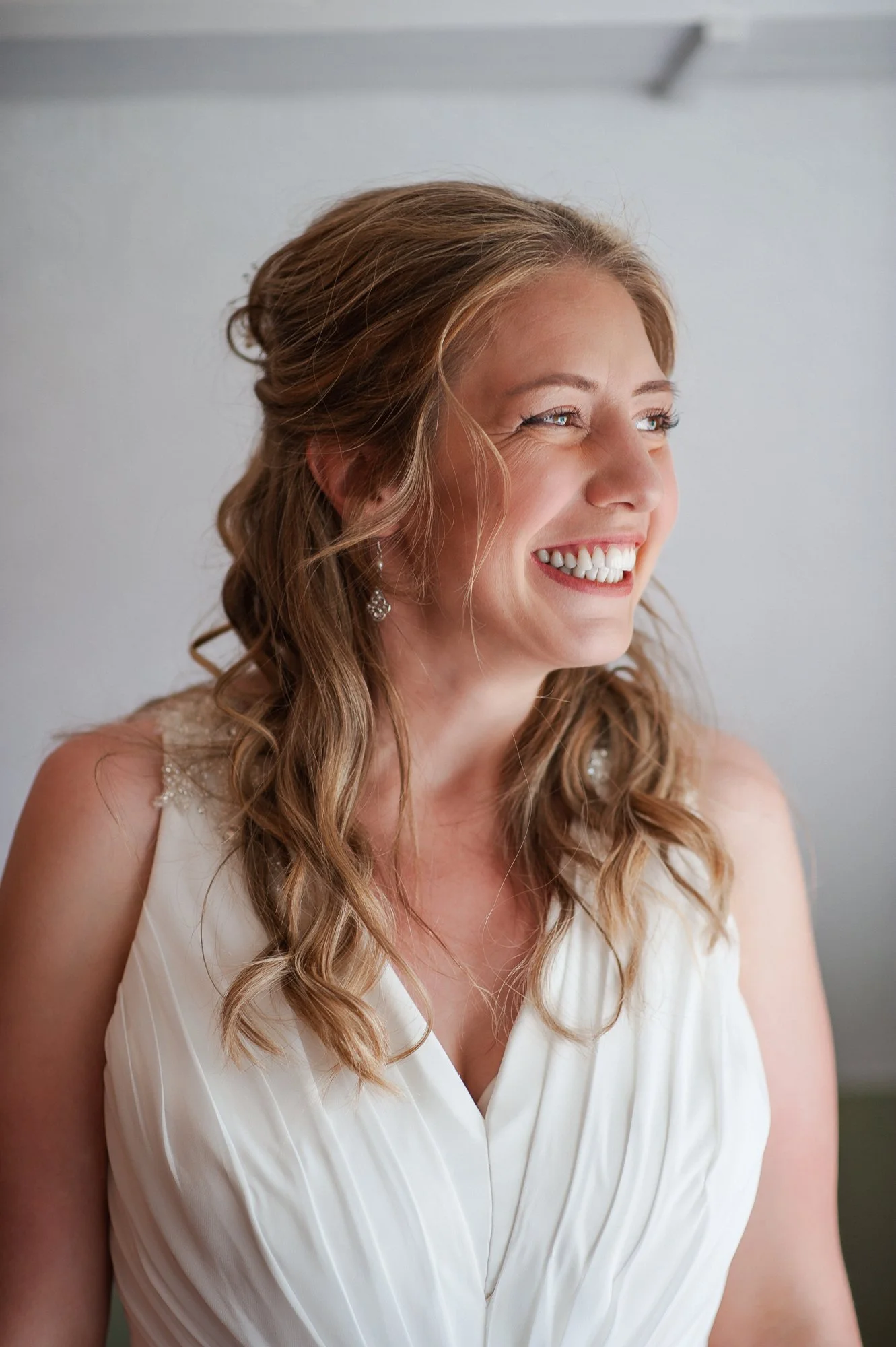 A smiling woman with wavy, shoulder-length hair, wearing a white sleeveless dress and diamond earrings, looking to the side.