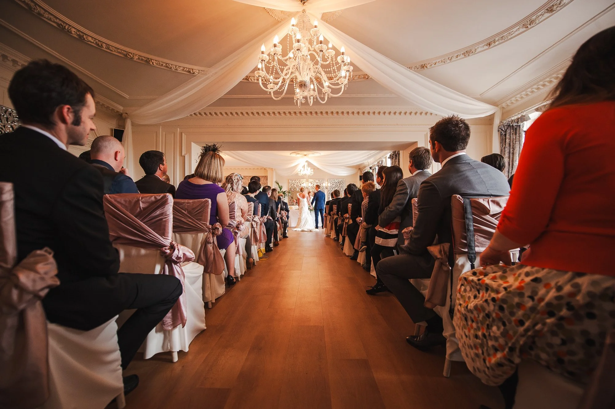 Wedding ceremony with guests seated on both sides, facing the bride and groom at the altar, decorated with chandeliers and elegant drapery.