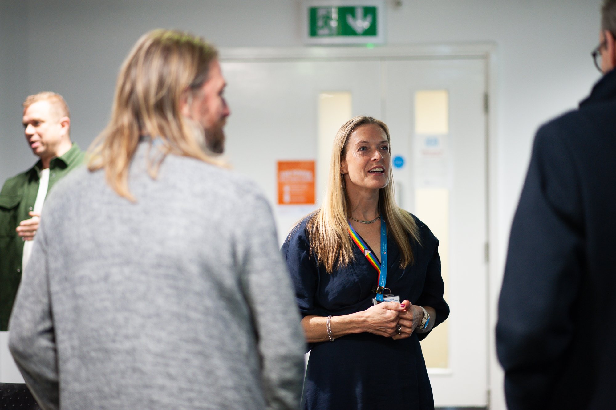 A group of four people, three men and one woman, engaged in a conversation indoors, with a green emergency exit sign above the door in the background.