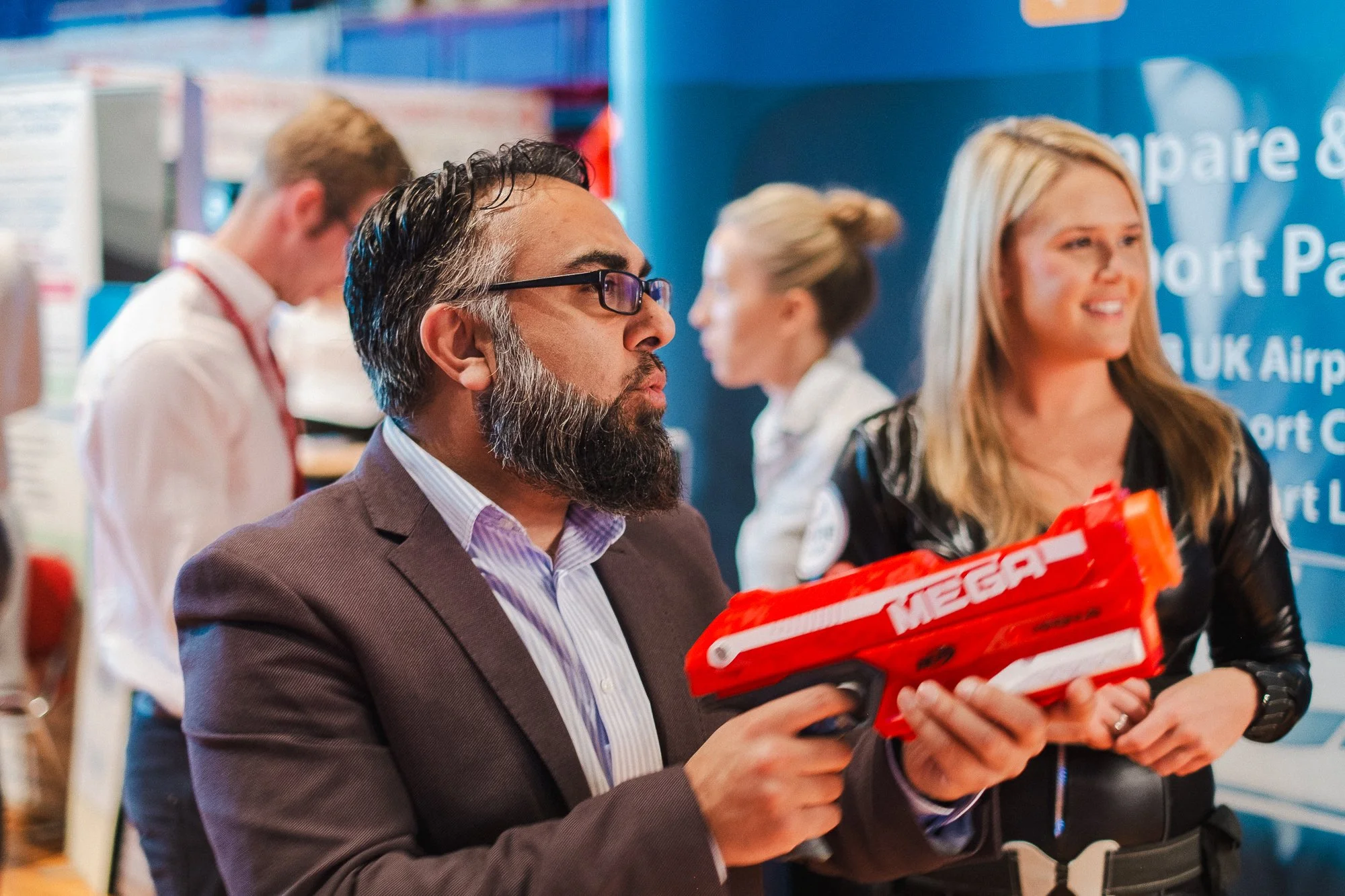 A man with glasses, a beard, wearing a suit jacket and shirt, holds a red toy gun and appears to be aiming or inspecting it, standing in an indoor setting with other people in the background.