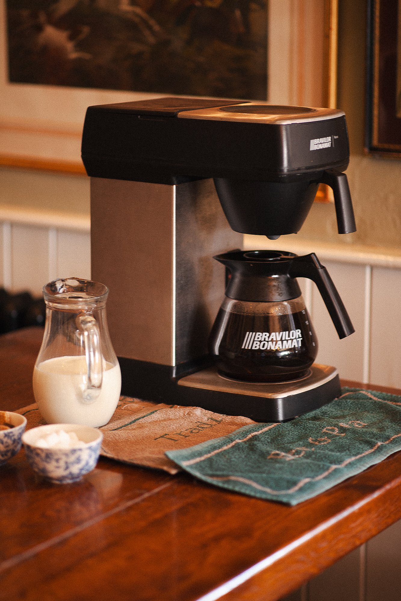 A coffee maker on a wooden table with a pitcher of milk and two small bowls, one containing powder, in a cozy room with framed pictures on the wall.