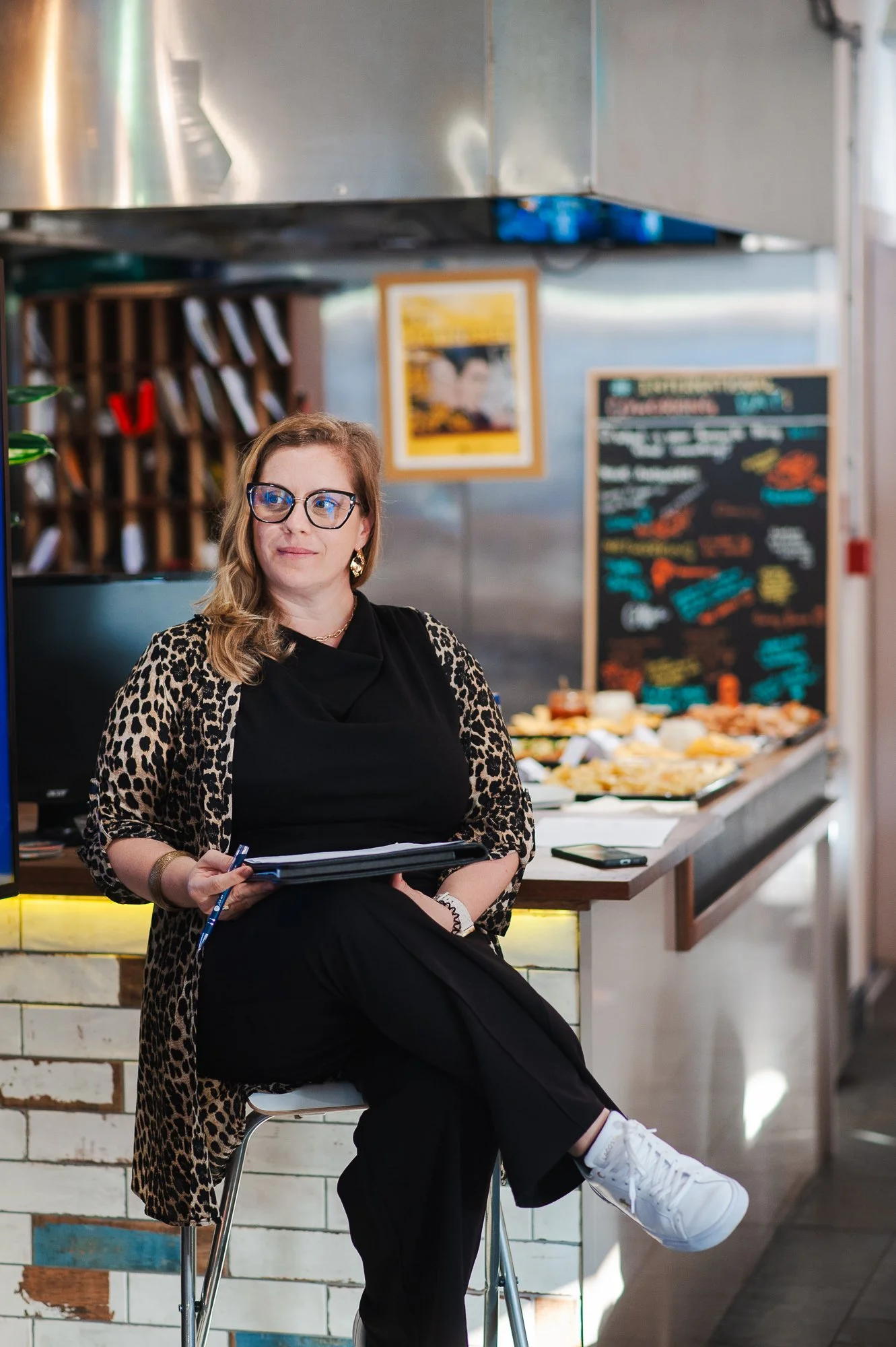 A woman with glasses sits on a stool in a restaurant or cafe, holding a folder or notepad, with a food display and a chalkboard menu in the background.