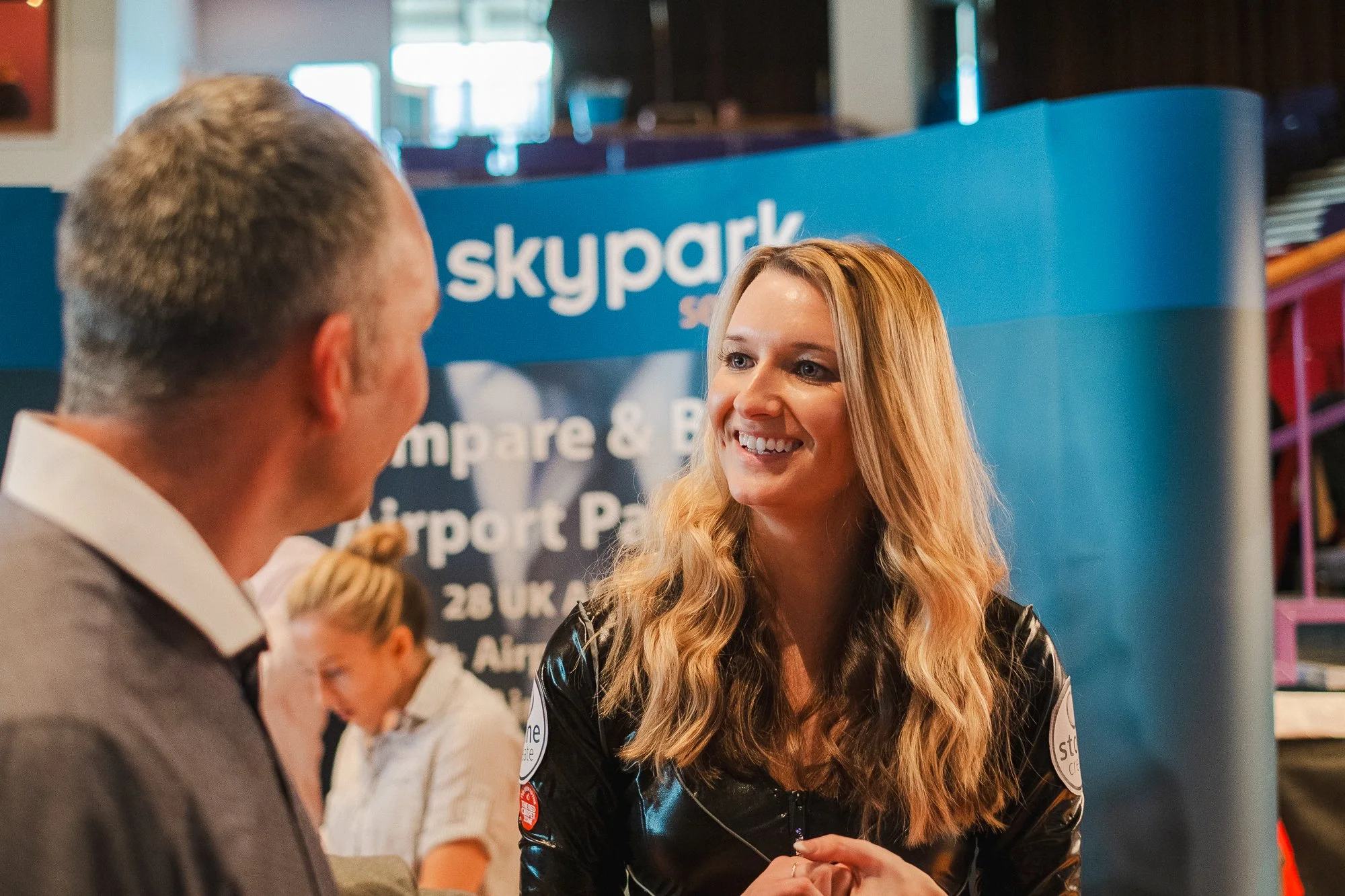 A woman with wavy blonde hair smiling and talking to a man with gray hair at an event with a blue Skypark banner in the background.