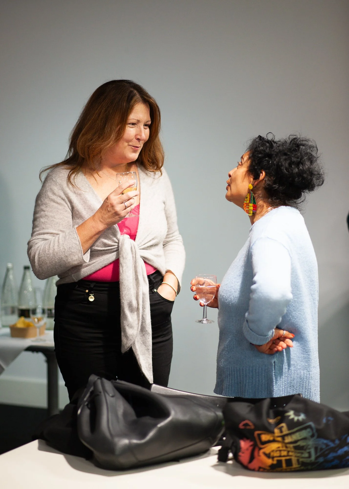Two women engaged in conversation, holding glasses of wine, with a table in the background that has bottles and snacks.