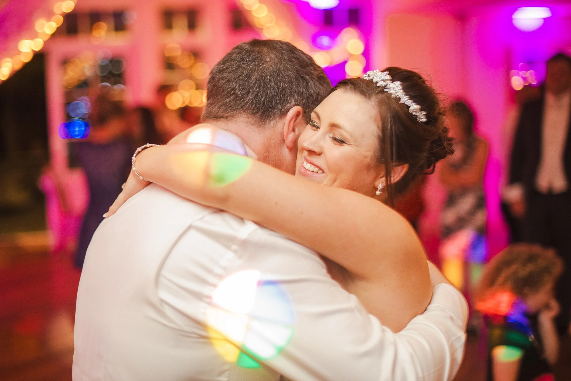 A woman in a wedding dress and a man in a white shirt share a joyful hug at a wedding reception, with colorful lighting and other guests in the background.