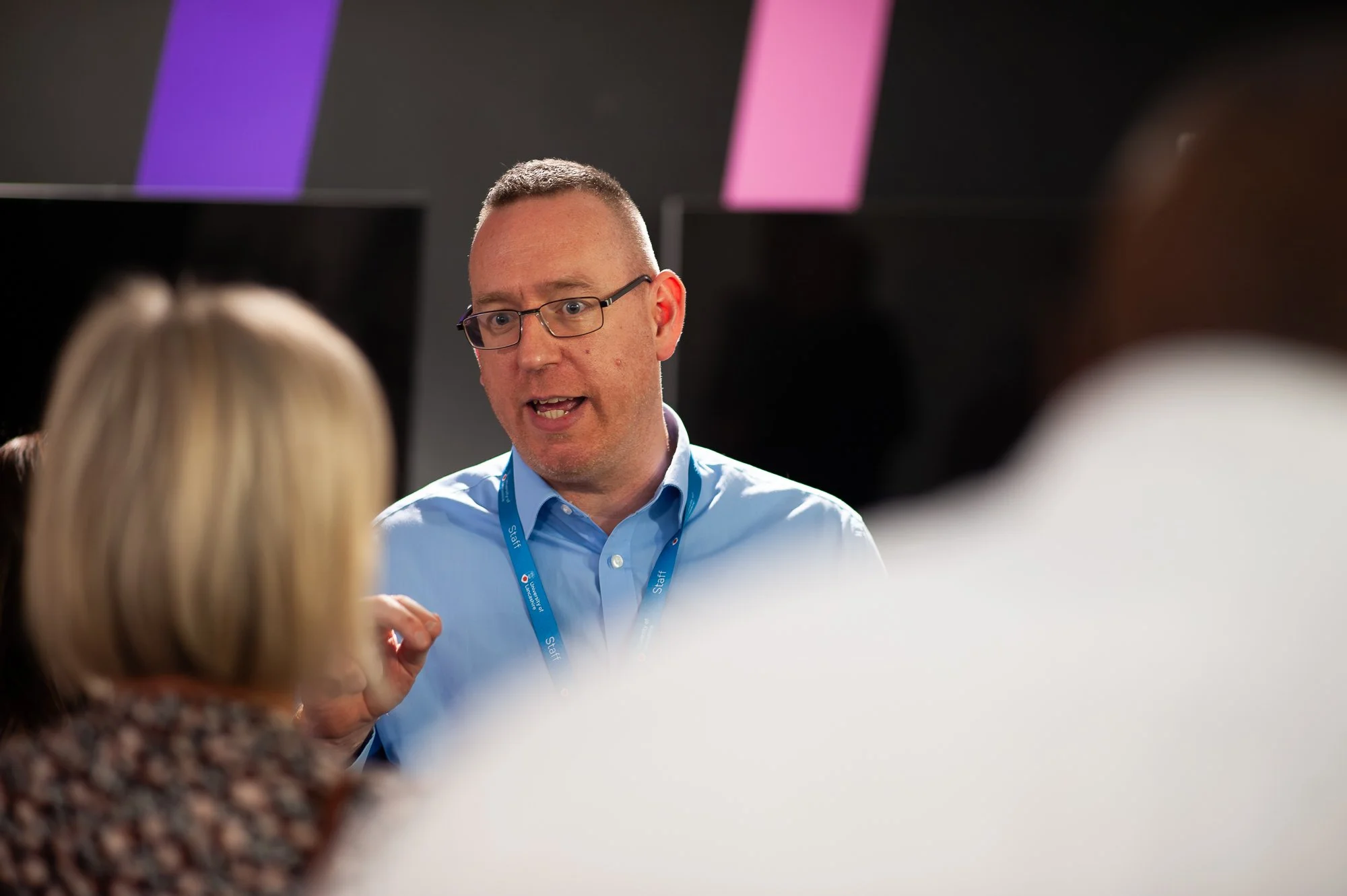 Man in glasses and blue shirt speaking, surrounded by people, in a professional setting with screens or panels in the background.