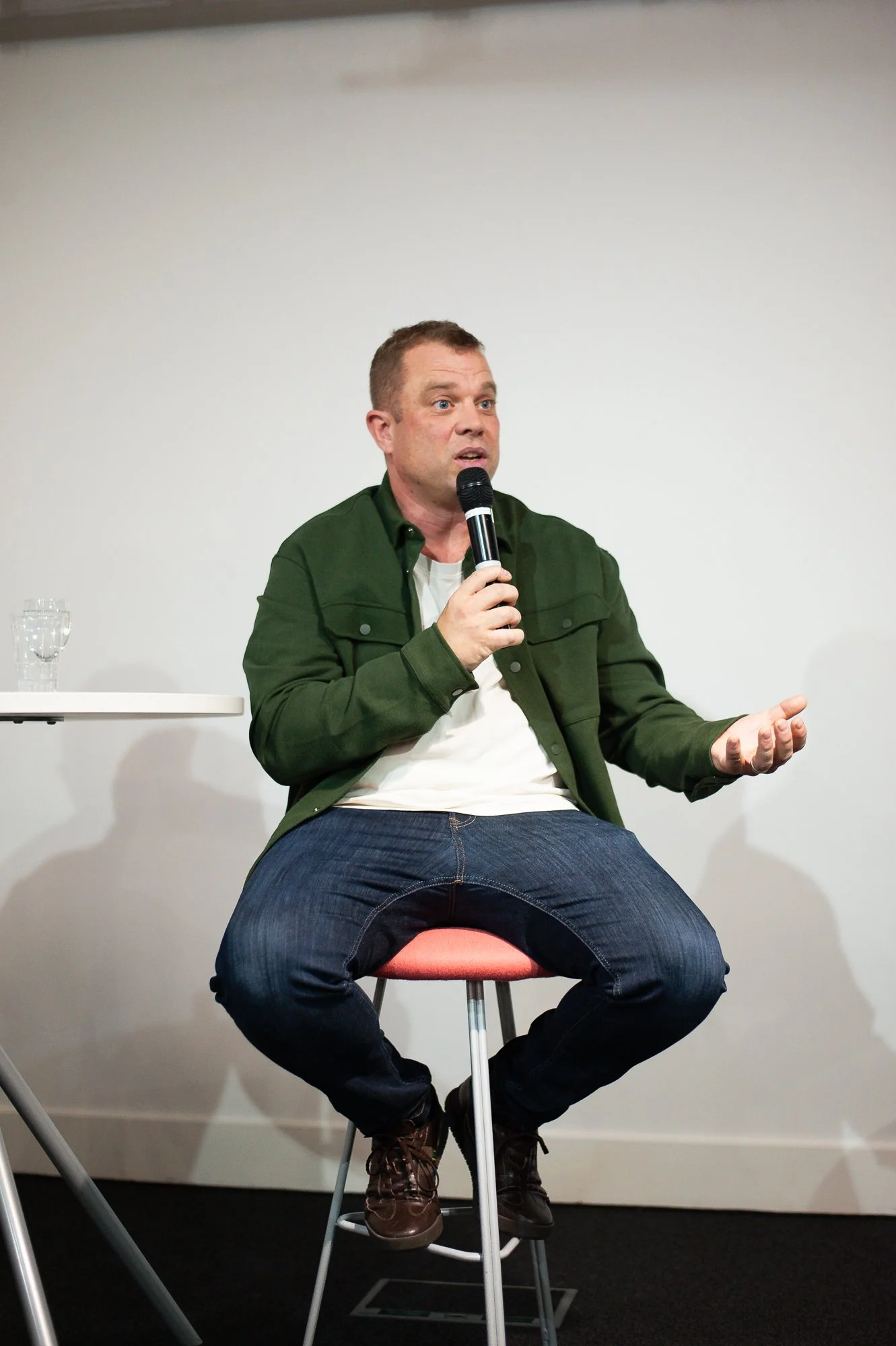 A man sitting on a stool, holding a microphone, speaking during an event or discussion.