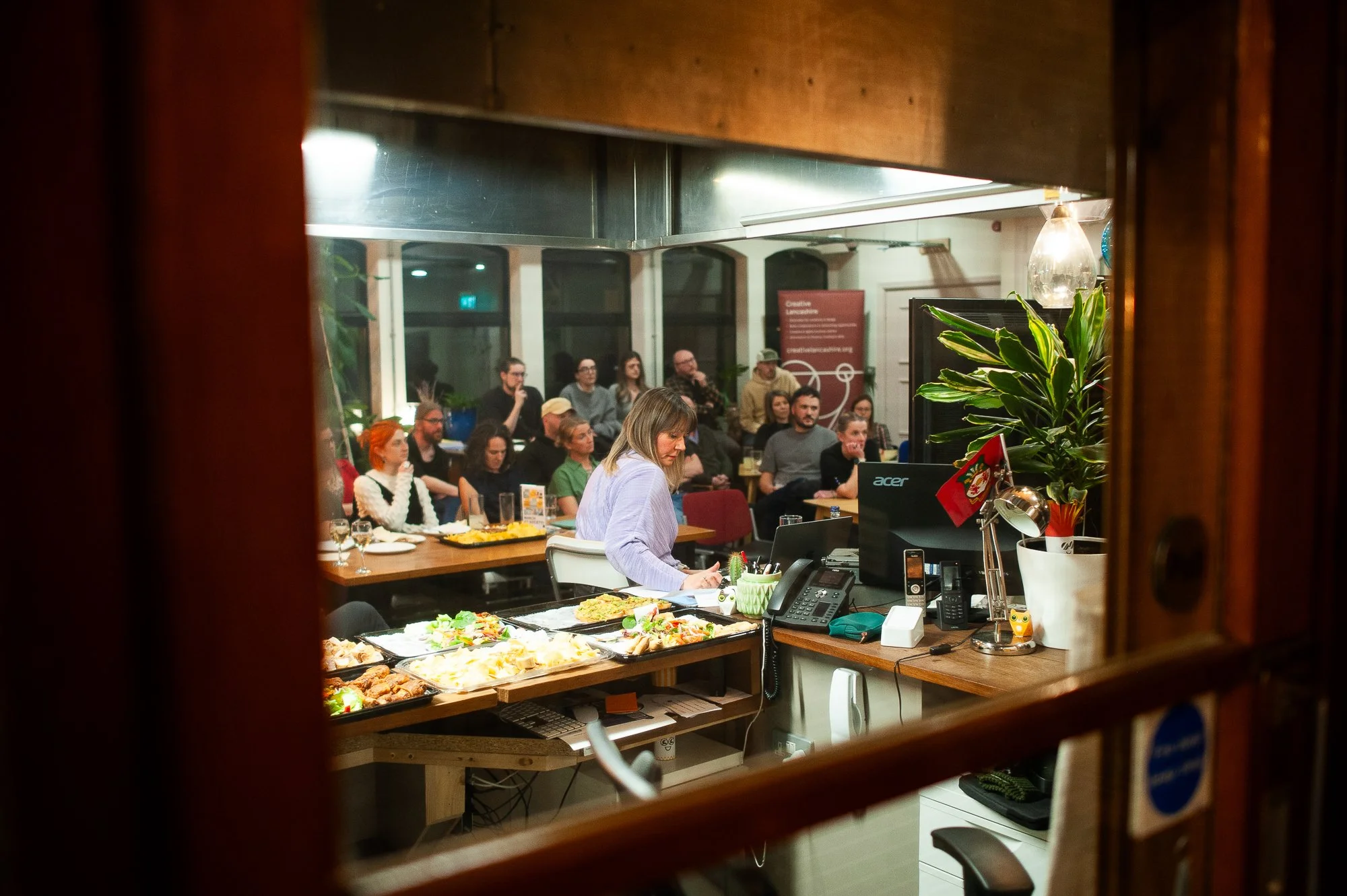 View through a window showing a woman working at a desk with food, and an audience seated in a room behind her during an event or presentation.