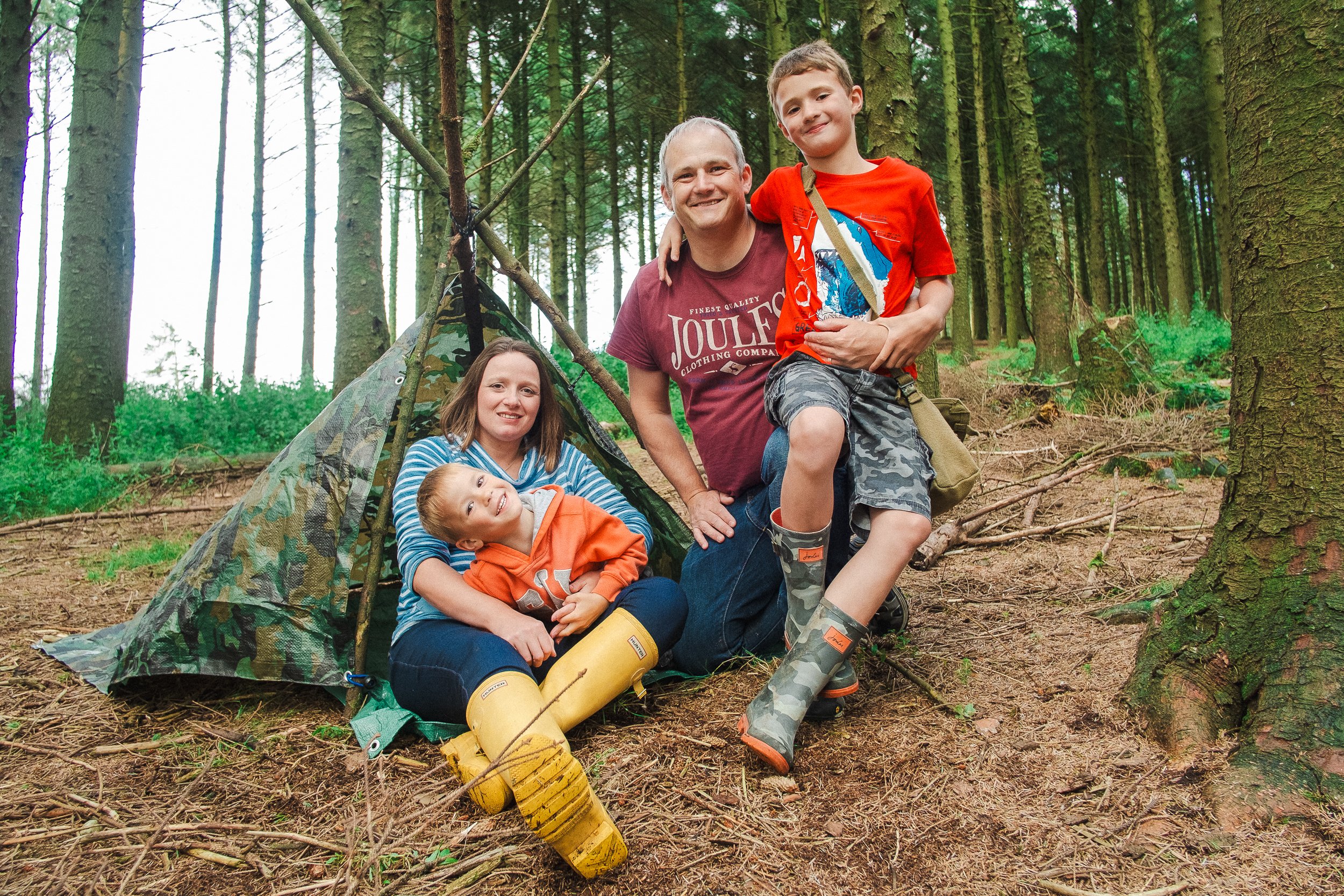 Family with two children posing in a forest next to a tent made of branches and camo tarp.