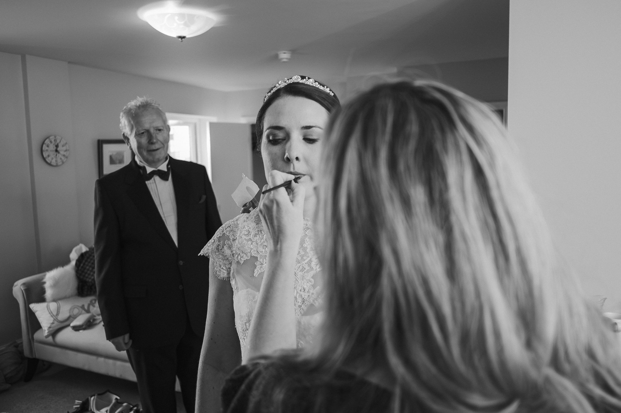 A bride wearing a lace wedding dress and a tiara getting her makeup done by a woman with long blonde hair, while a man in a tuxedo watches in the background in a living room.