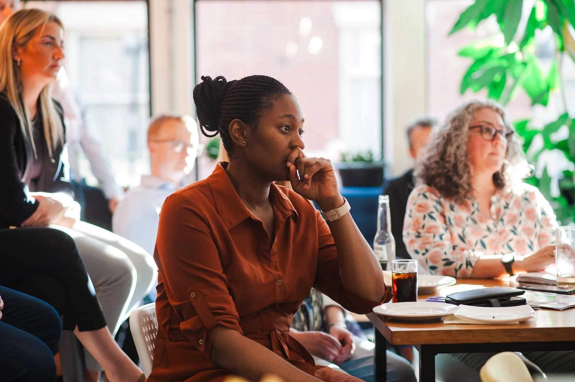 A woman with dark hair in a bun, wearing an orange-brown shirt, sitting at a table in a meeting room, listening attentively with her hand on her face.