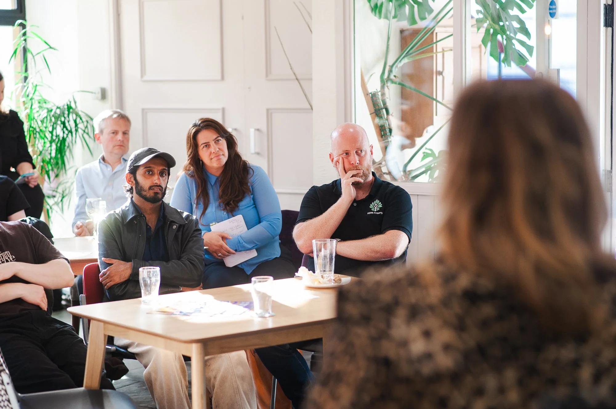 People sitting at a table in a room during a meeting or presentation, with some attendees listening attentively and others taking notes, while a woman is speaking at the front.