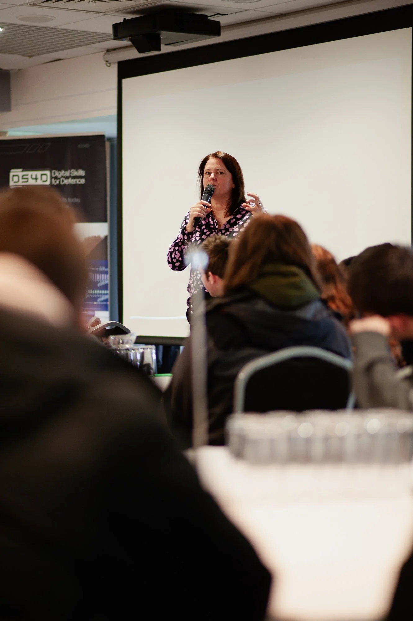 A woman presenting at a conference, standing in front of a large projection screen, talking into a microphone while gesturing with her hand. Audience members are seated and listening.