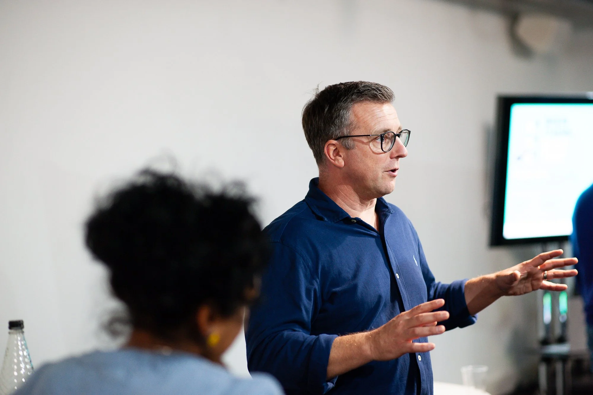 A man in a blue shirt and glasses is giving a presentation or speech in a classroom or conference room. Someone with curly hair and yellow earrings is partially visible in the foreground. A screen is visible in the background.