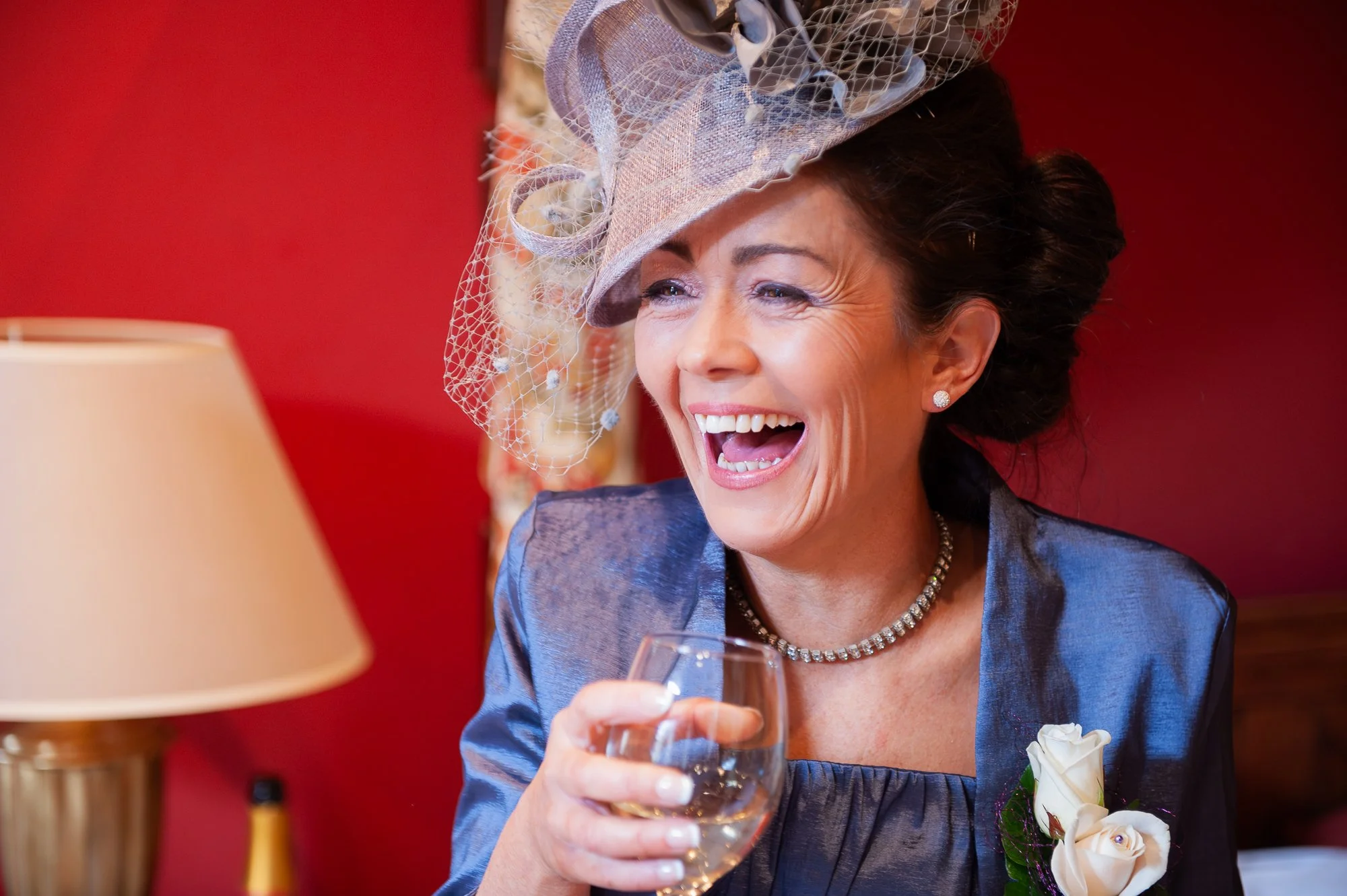 A woman laughing while holding a glass of white wine, dressed elegantly with a pearl necklace, earrings, and a fancy hat with netting, in a room with red walls and a lamp.