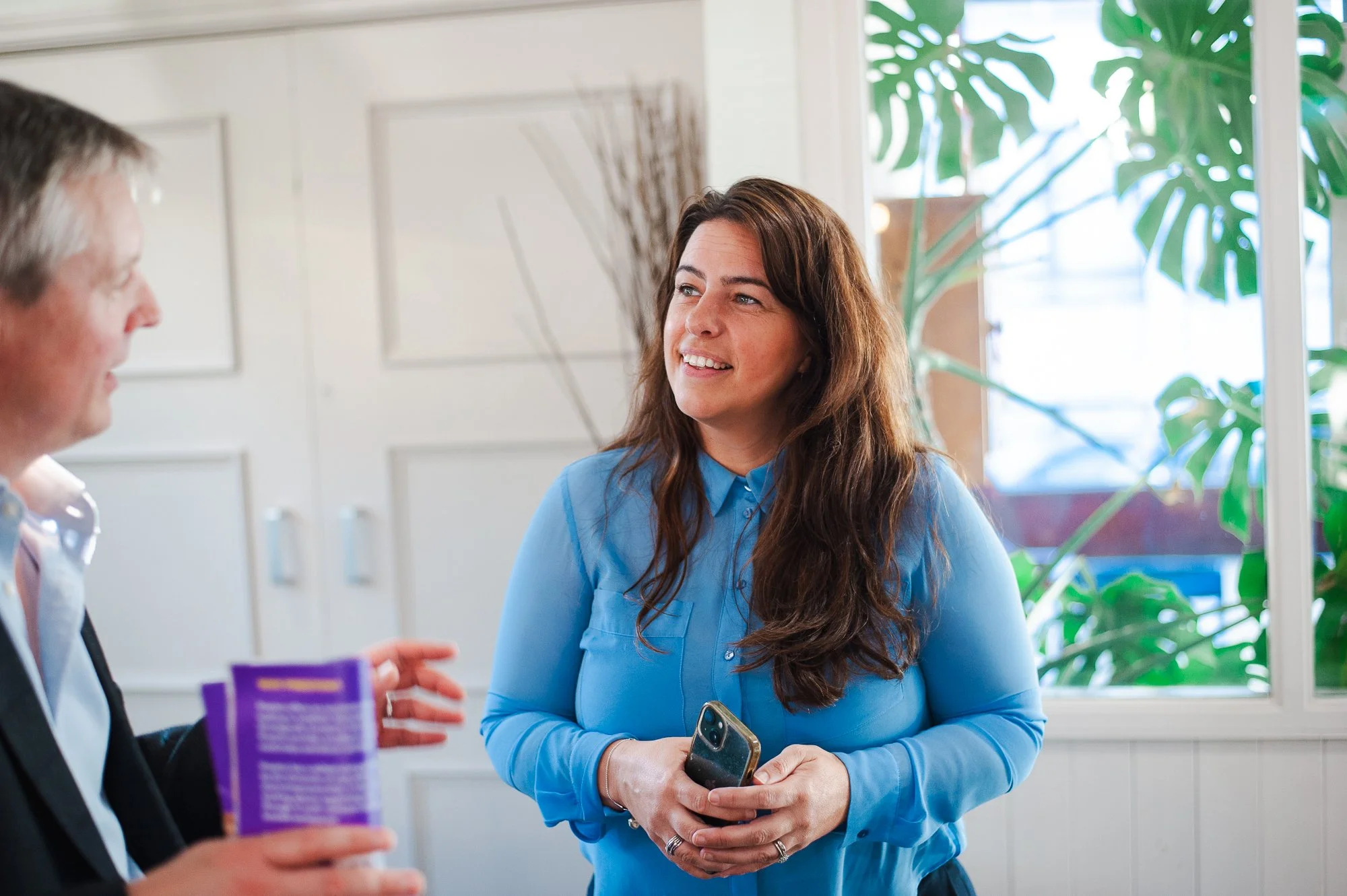 Two people having a conversation indoors, with a woman in a light blue shirt holding a phone, smiling, and a man with gray hair pointing and holding a purple item.