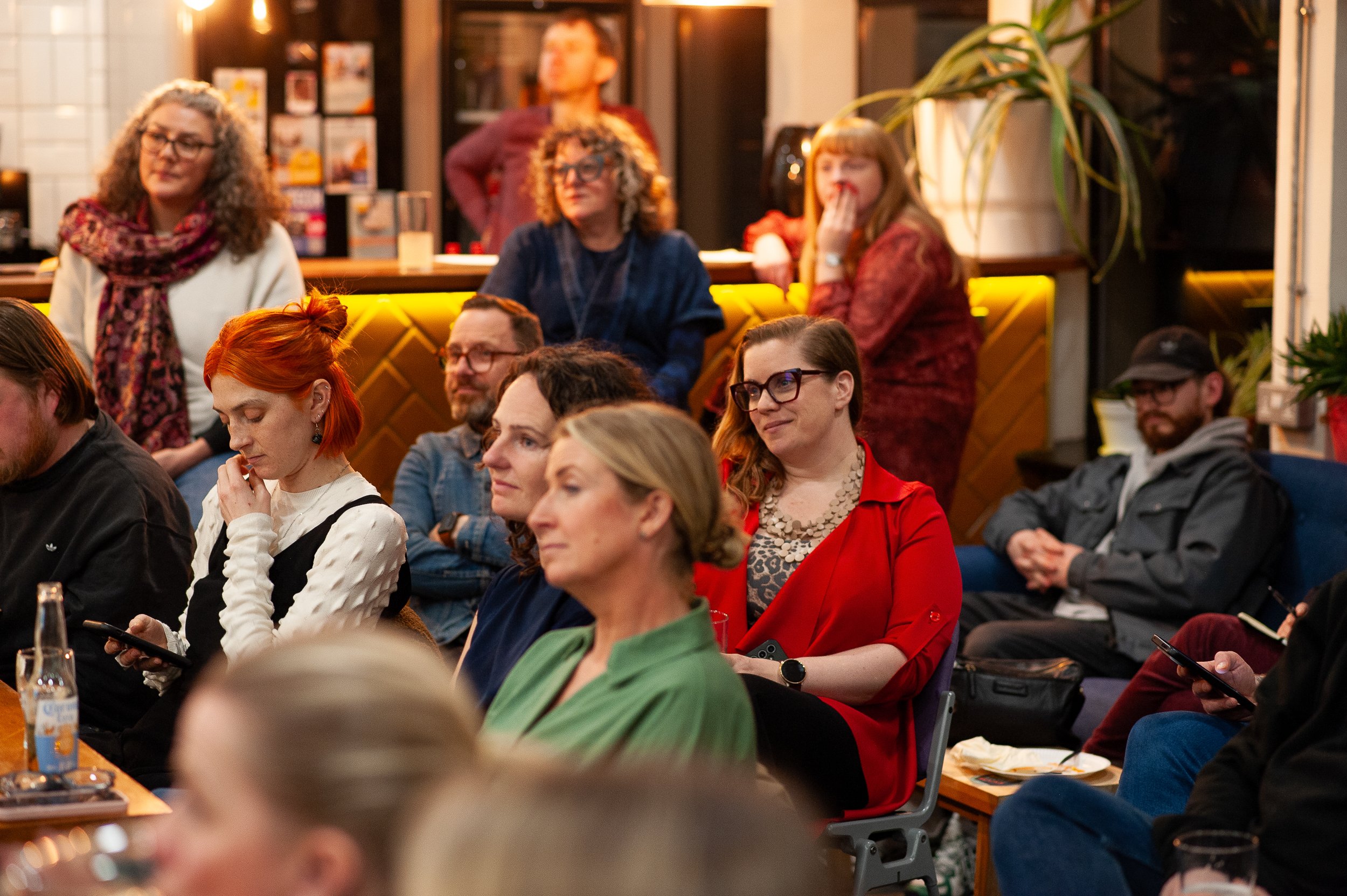 Audience attentively watching a presentation in a cozy indoor setting with plants and warm lighting.