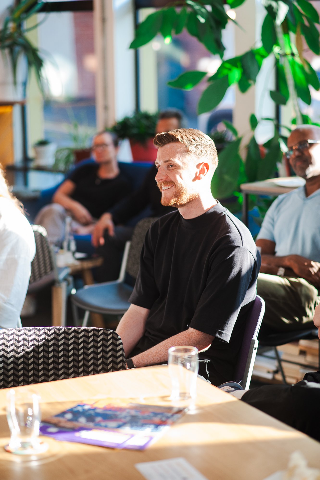 A group of people attending a meeting or presentation in a well-lit room with large windows and green plants. The focus is on a young man with light brown hair and a beard, smiling, wearing a black t-shirt, sitting at a table.
